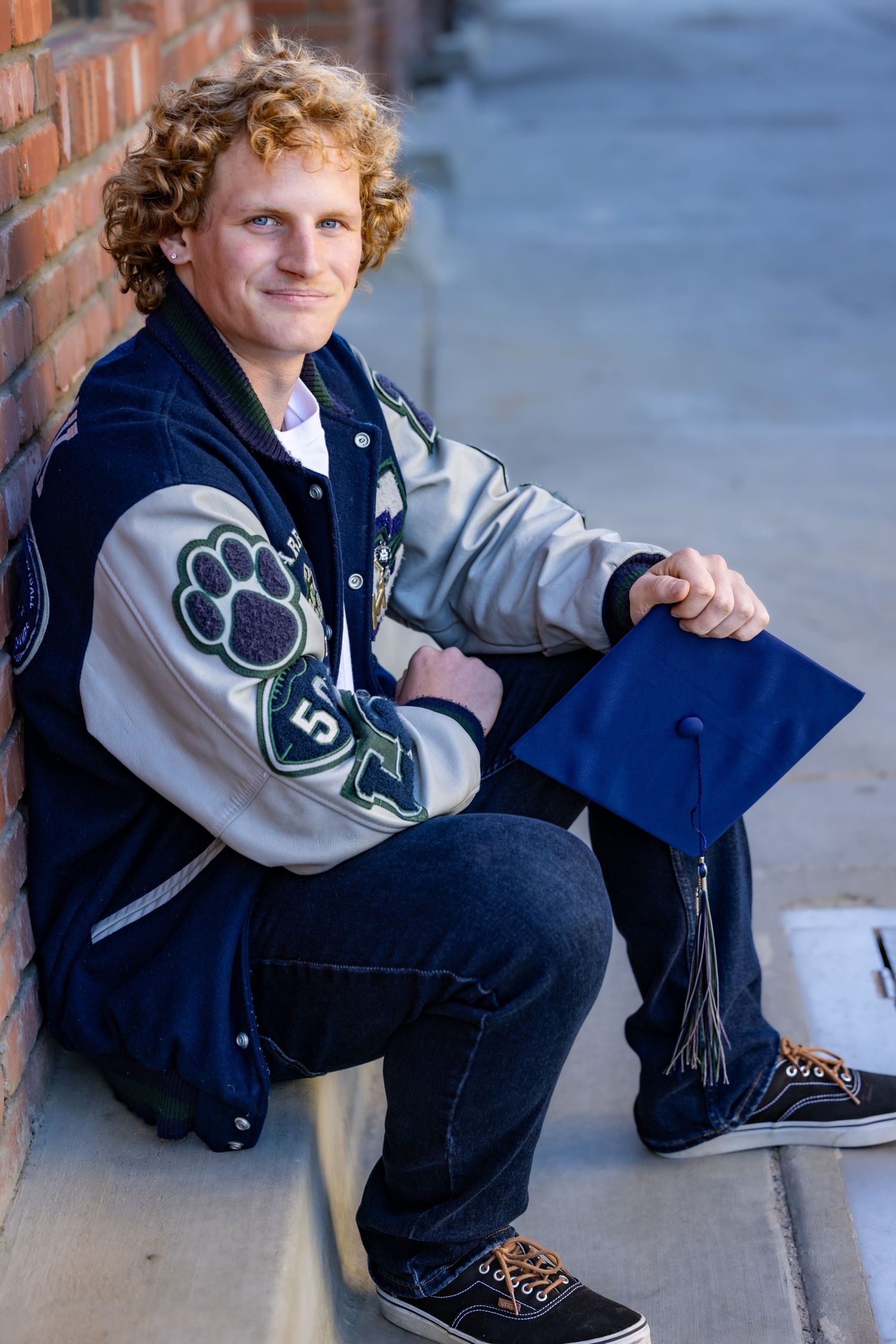 Person wearing a navy and beige varsity jacket sits casually against a brick wall in an outdoor setting.
