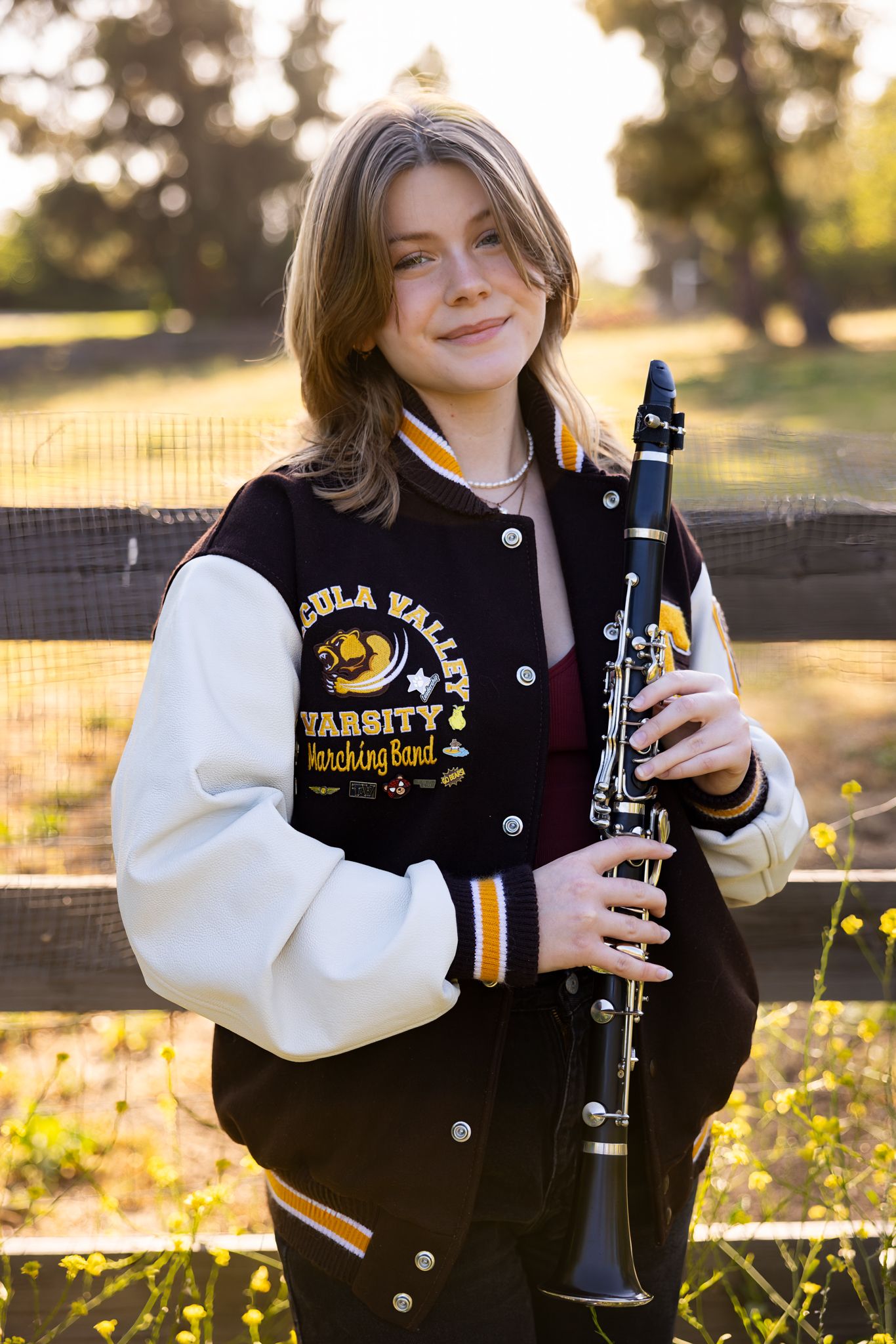 Student in varsity letterman jacket holds clarinet in golden afternoon sunlight during outdoor photoshoot.