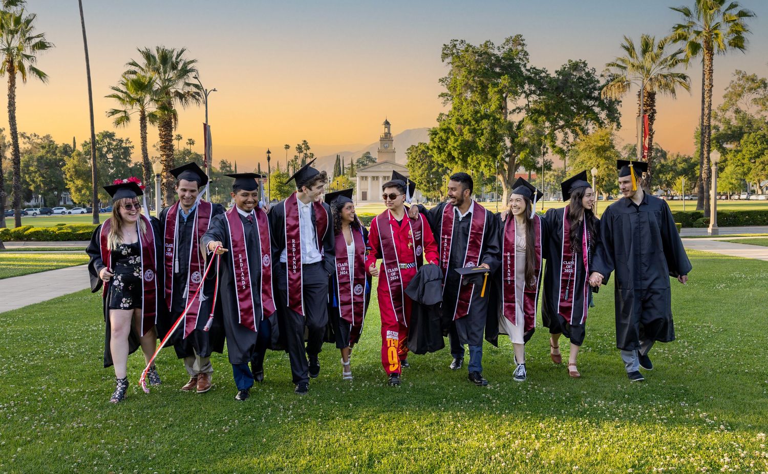 Graduates in red and black commencement robes stand together on a grassy lawn with palm trees at sunset.