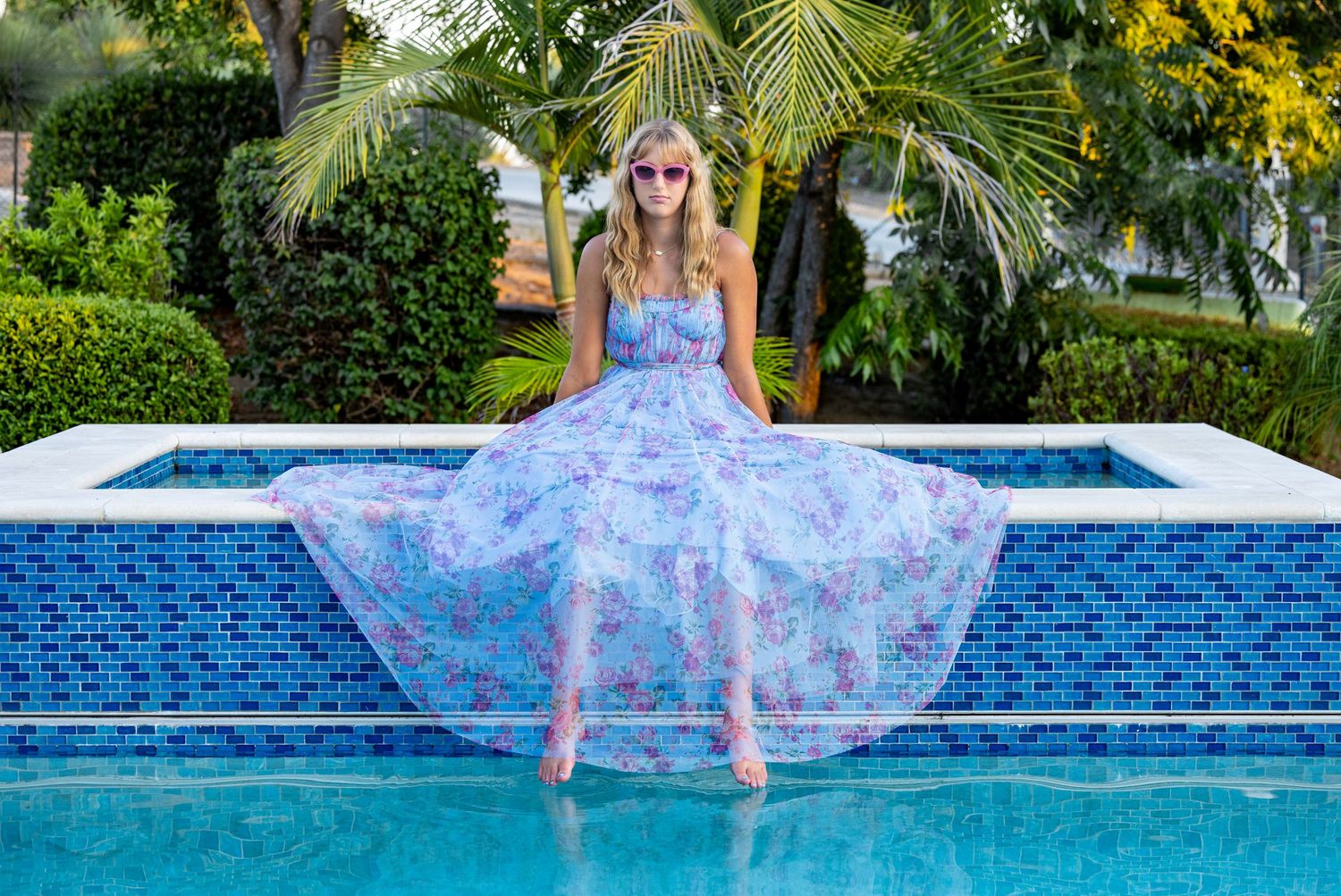 A flowy blue floral dress billows gracefully over a swimming pool with mosaic tiles in a tropical setting.