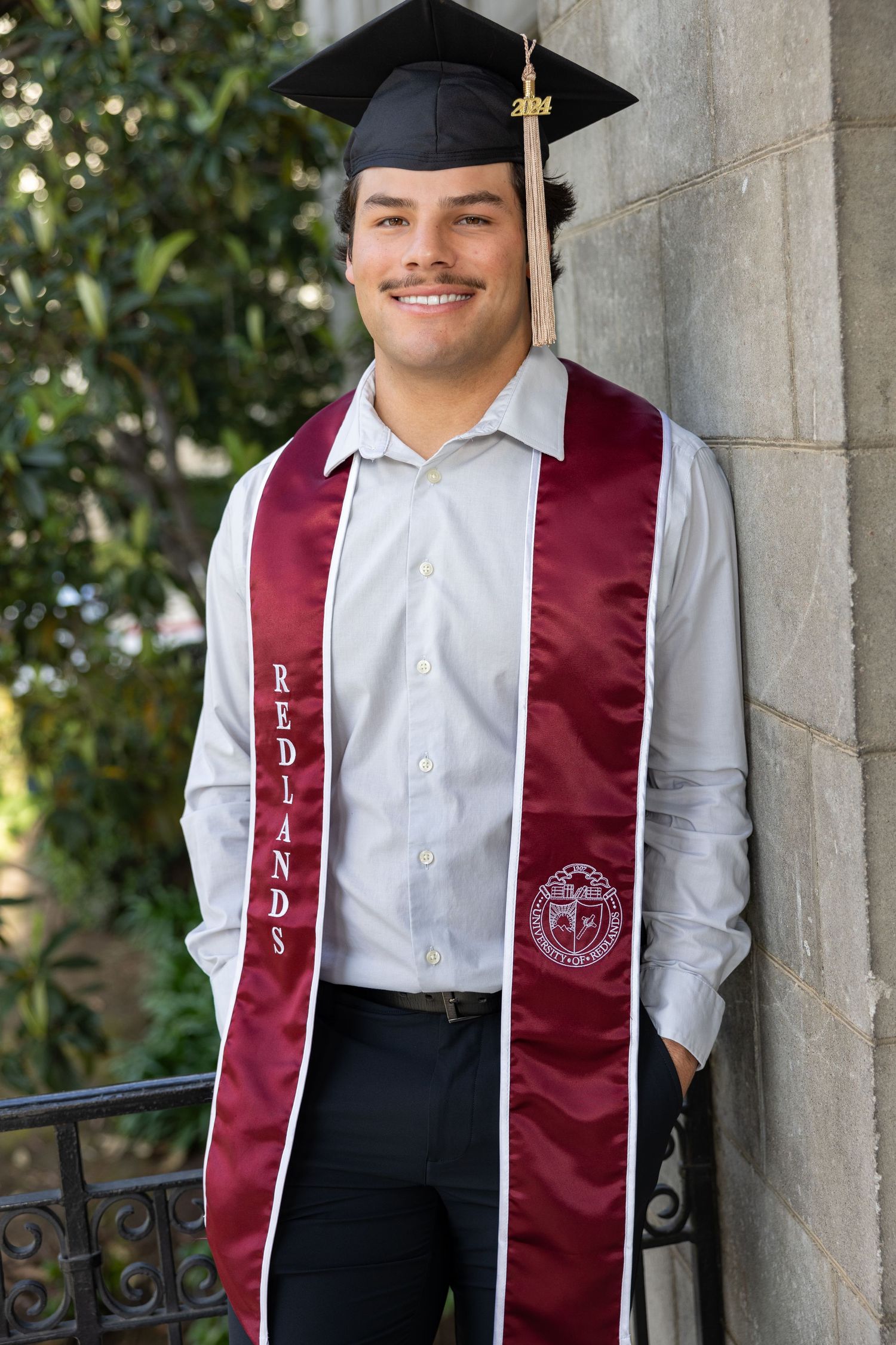 Graduate wearing maroon stole and graduation cap poses in front of stone columns during commencement ceremony.