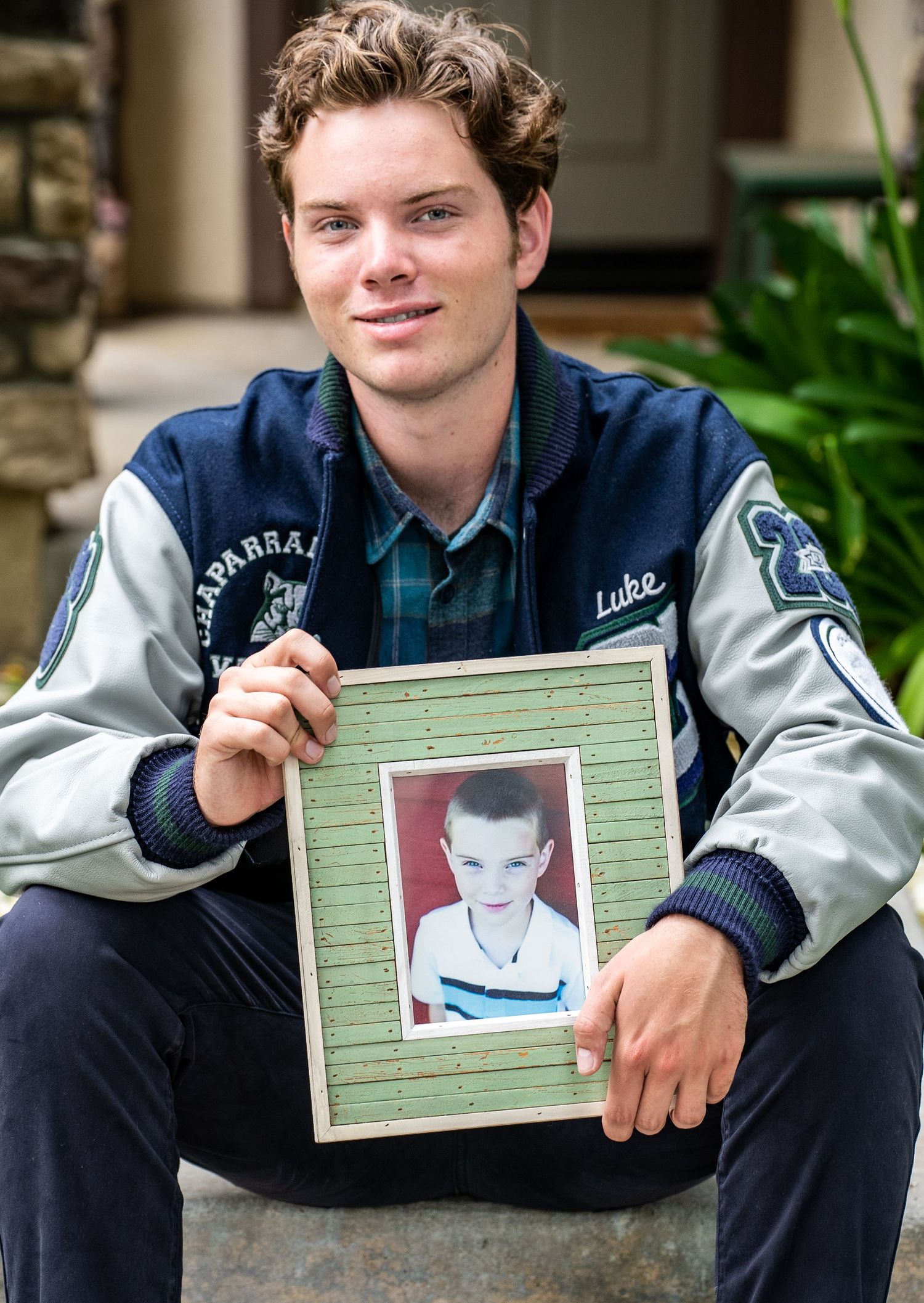 A person in a varsity jacket holds a green picture frame while sitting outdoors.
