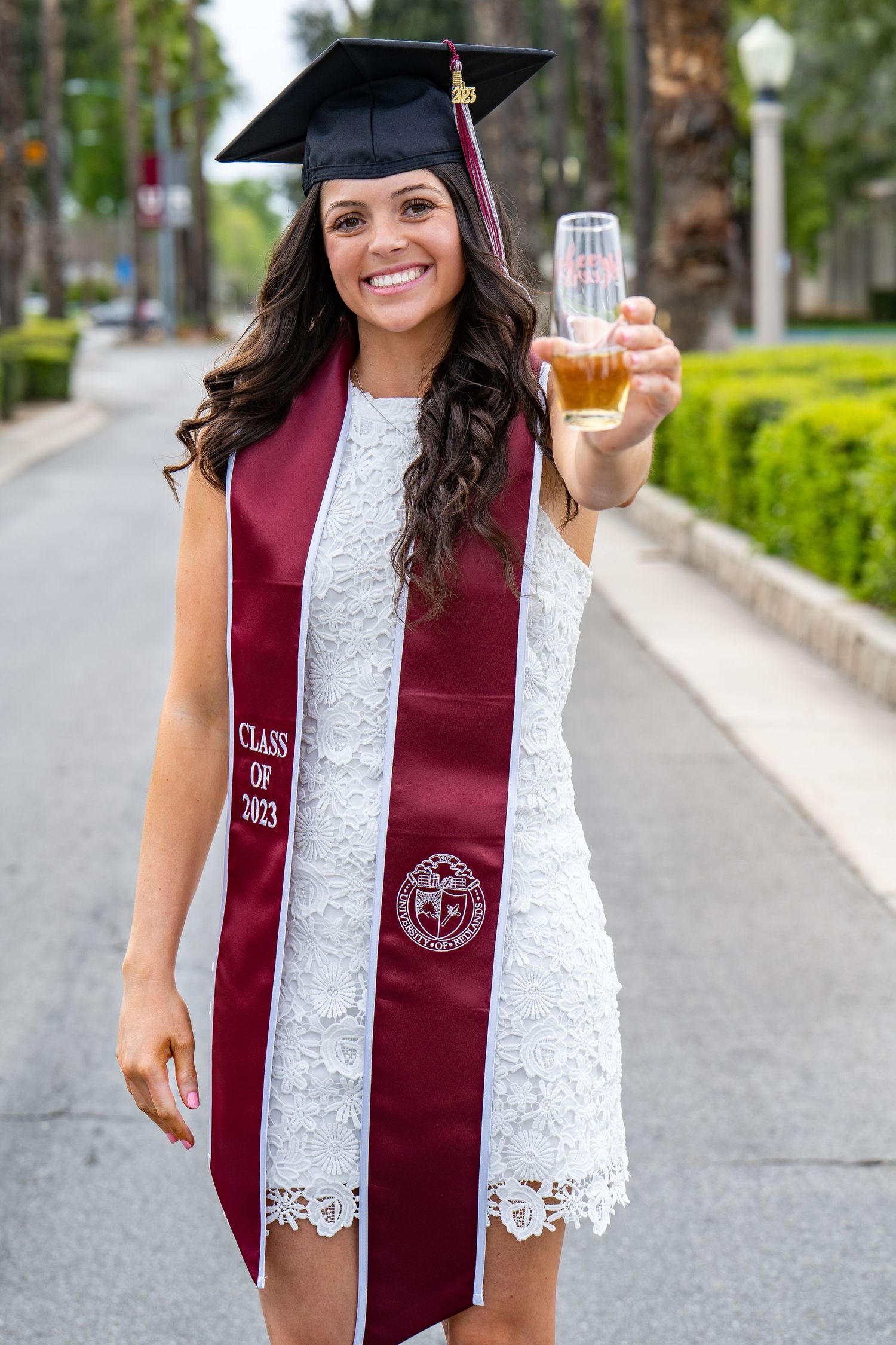 Graduate in white lace dress and burgundy graduation stole holds up a drink while celebrating on campus.