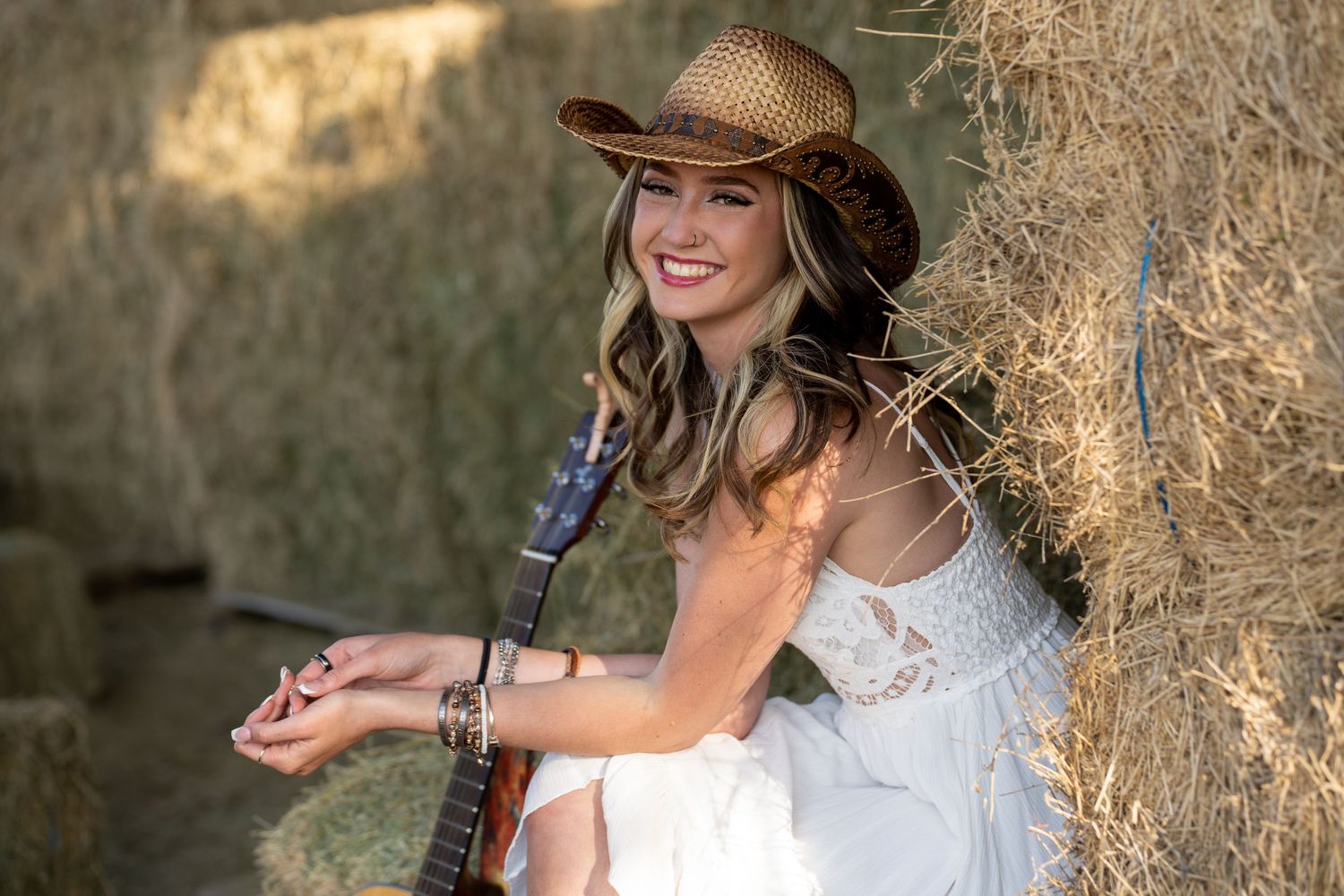 Woman in white dress and cowboy hat poses in rustic outdoor setting with hay bales in background.