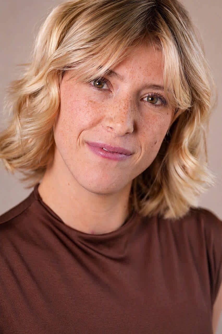 Smiling blonde woman with freckles wearing a brown t-shirt poses for a portrait photo against a neutral background.