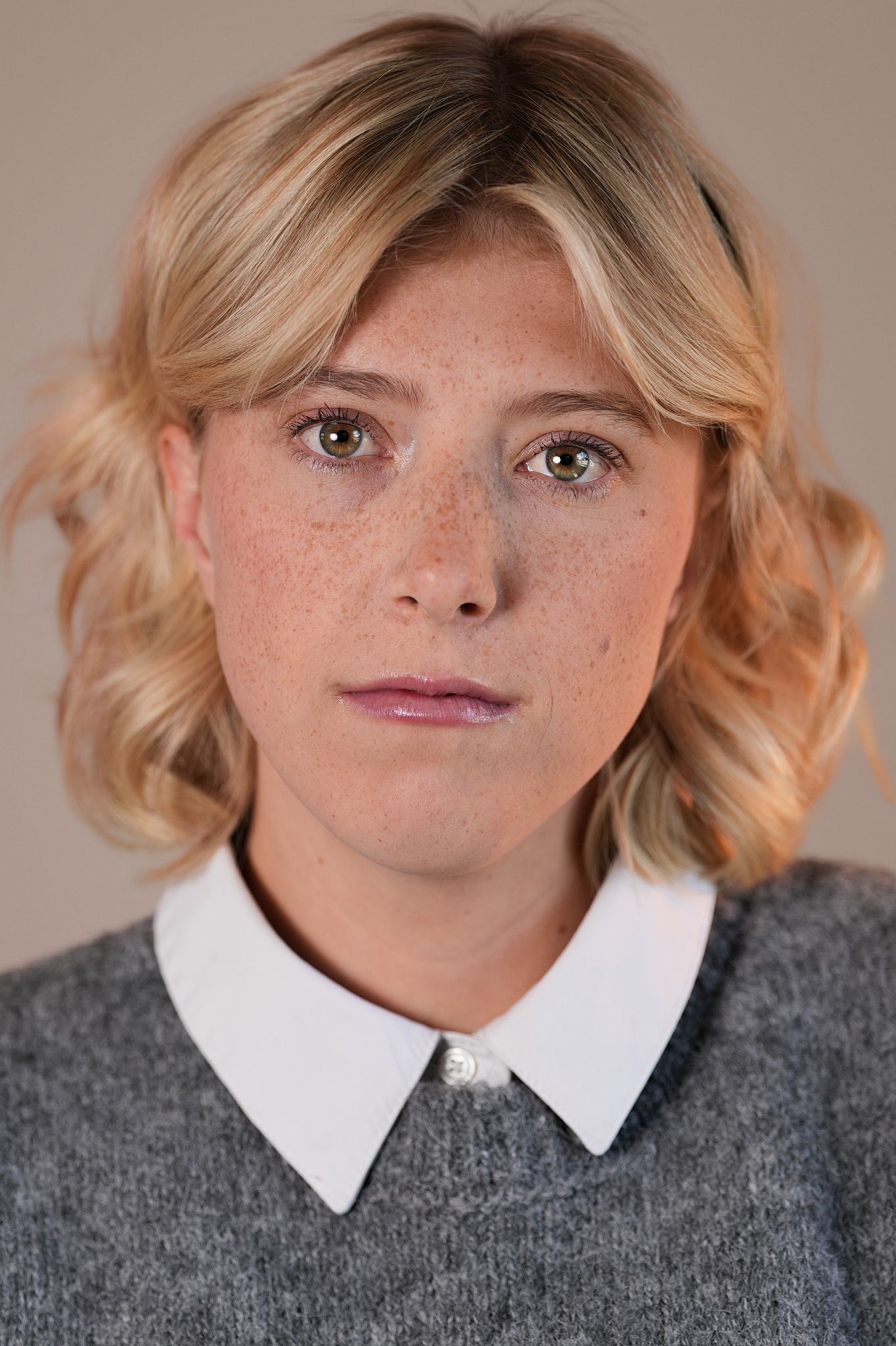 Young blonde woman with freckles and green eyes wearing a grey sweater with white collar, neutral expression.