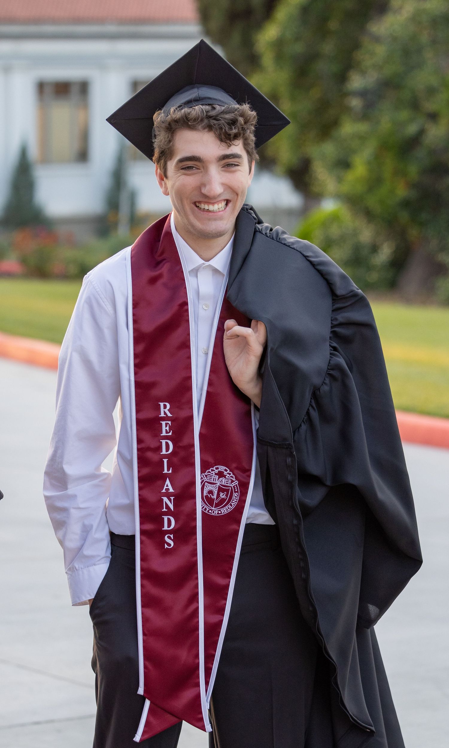 Graduate wearing maroon graduation stole and carrying black cap and gown walks outdoors on graduation day.