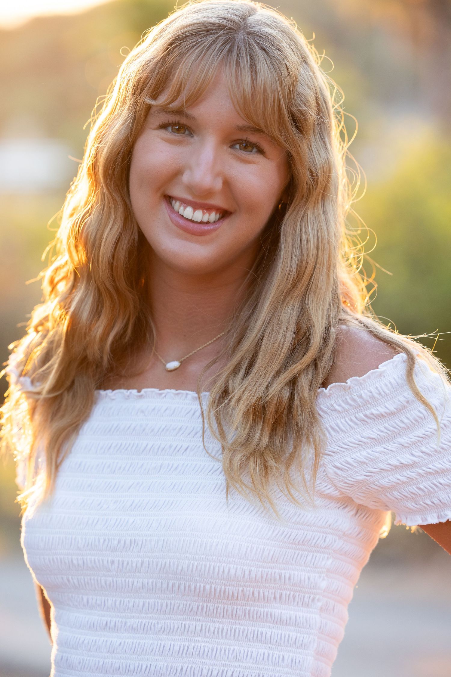 A person in a white lace top poses outdoors during golden hour with warm sunlit backlighting creating a glowing effect.