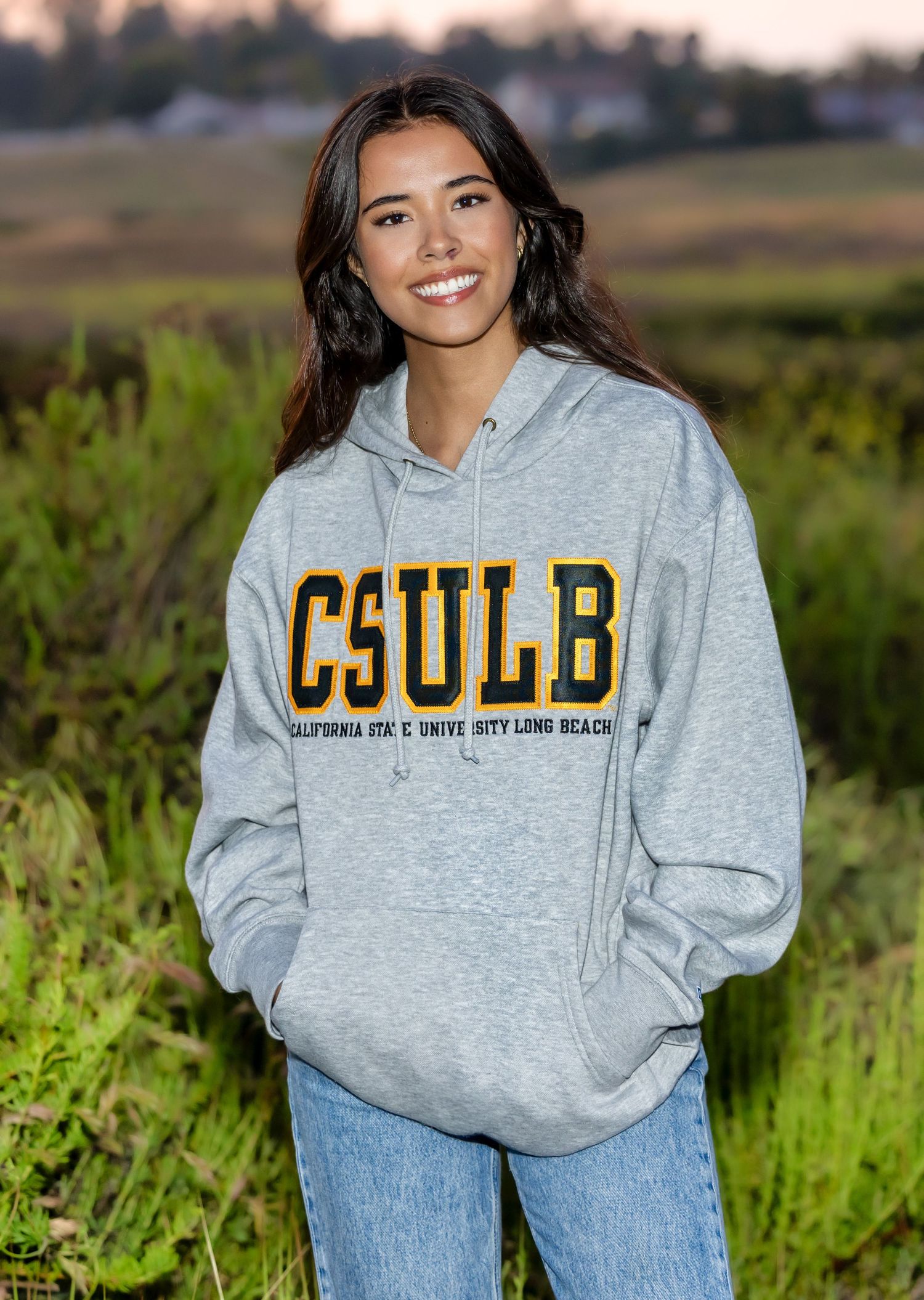 Person wearing a gray CSULB hoodie with gold lettering poses outdoors in grassy field at sunset.