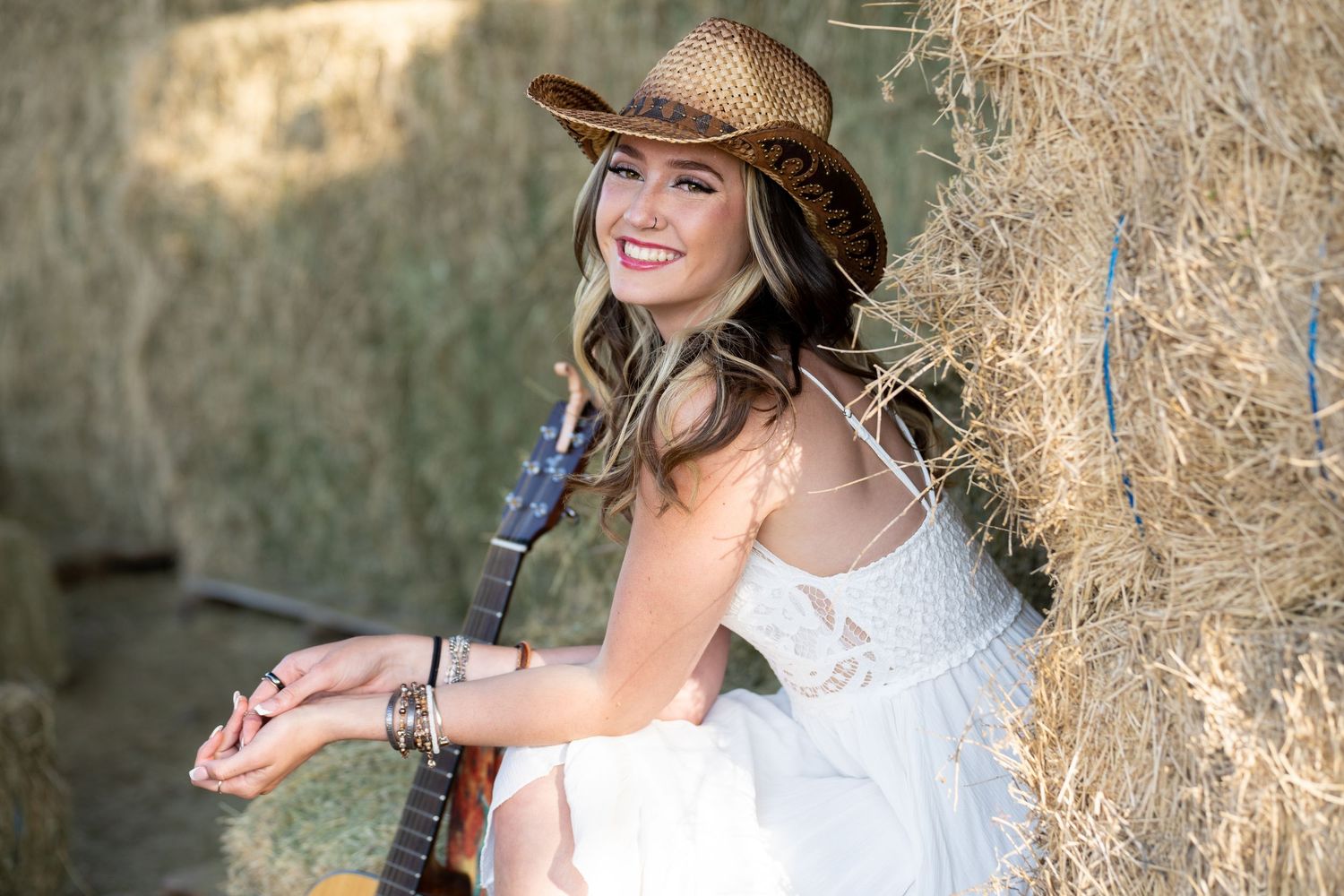 A woman in a white dress and cowboy hat smiles while sitting next to hay bales in a rustic setting.