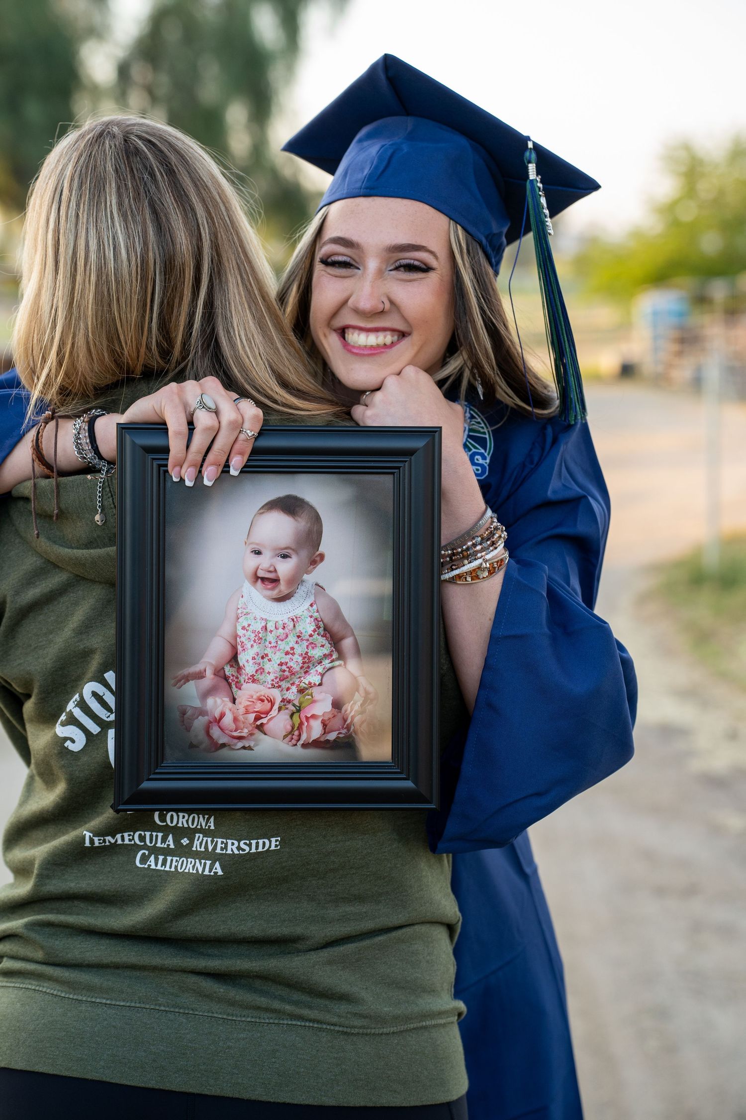 Graduate wearing olive sweatshirt and navy gown displays framed baby portrait outdoors.
