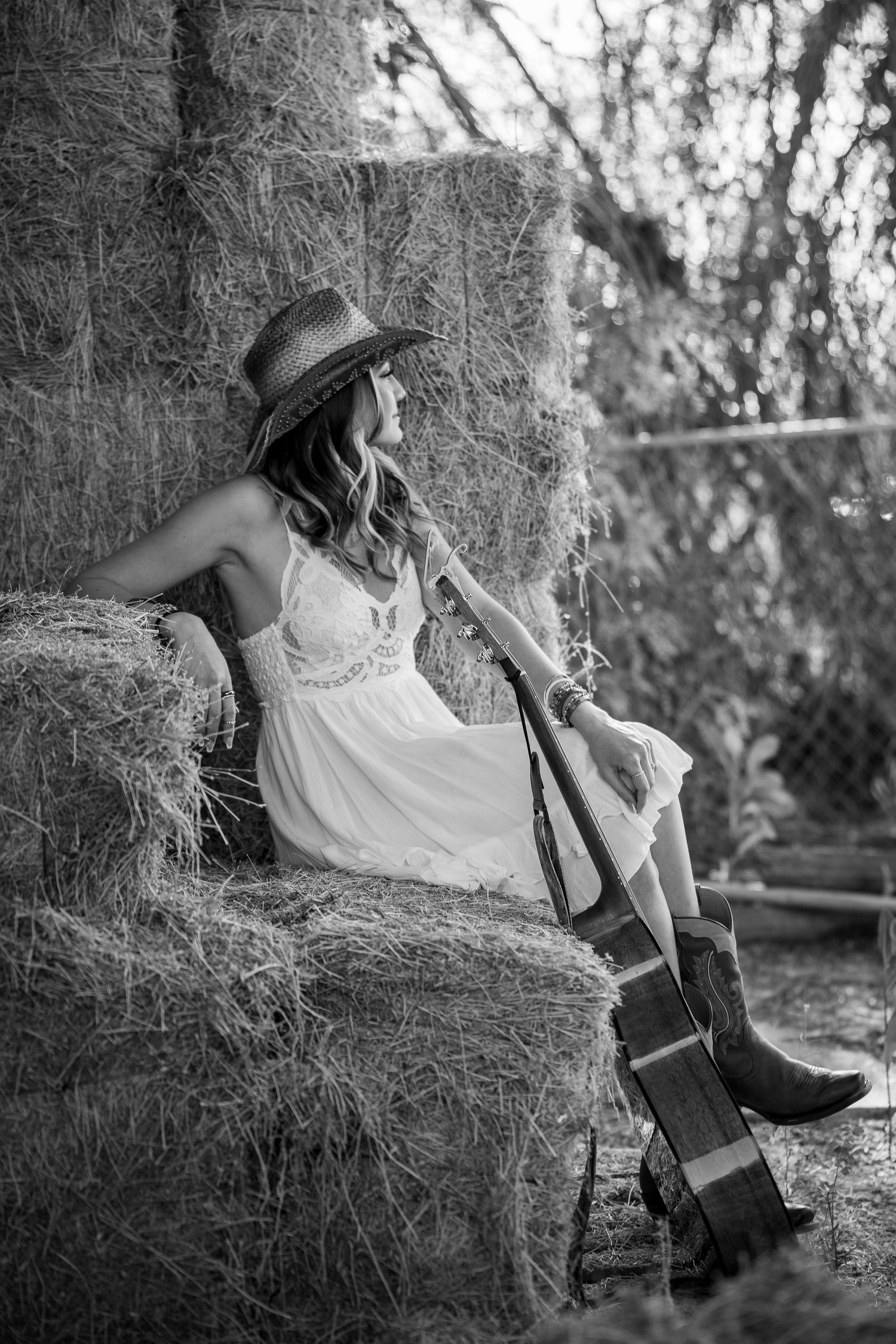 A person in a white dress and hat sits on Spanish moss while playing a violin in a black and white photograph.