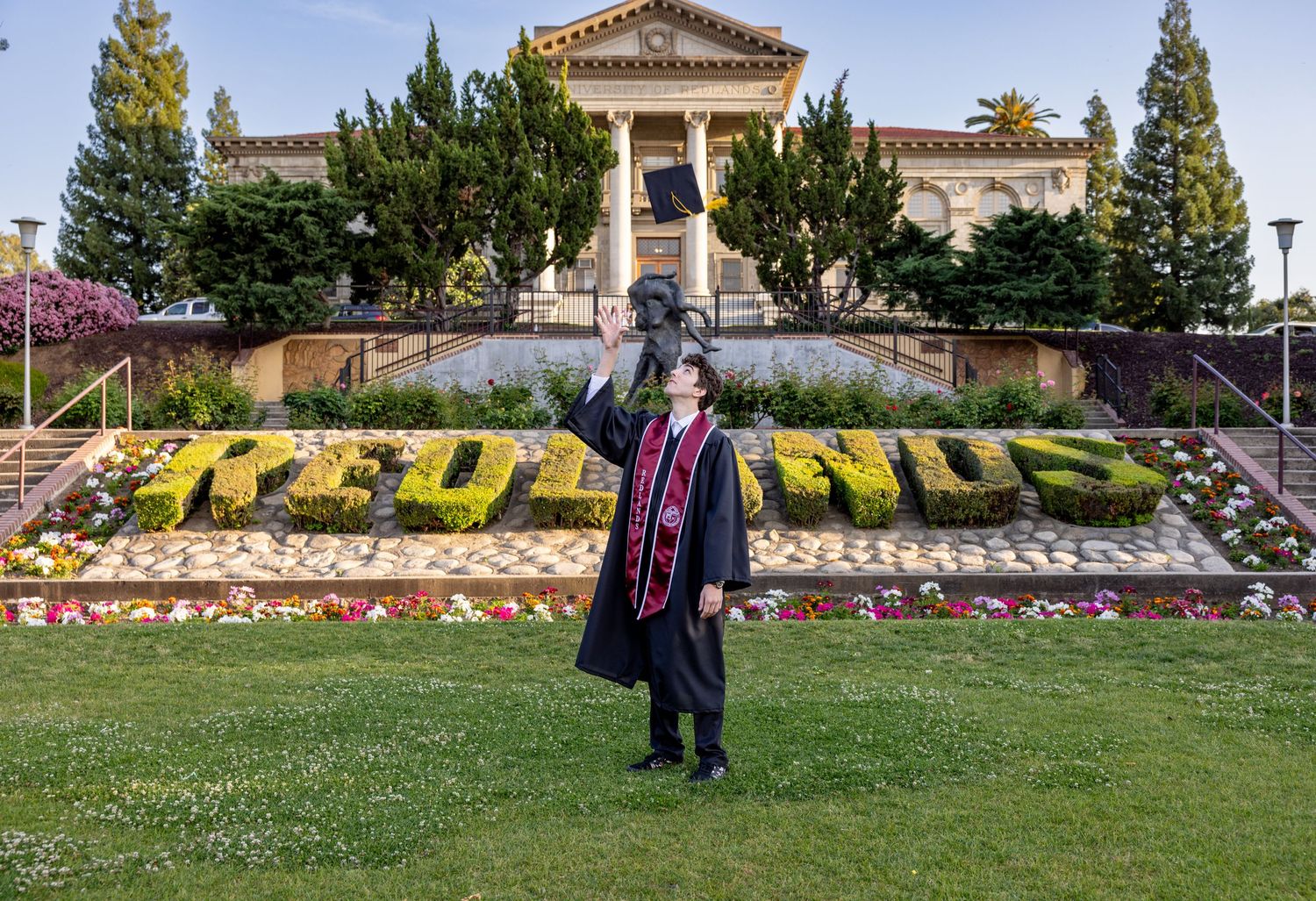 A panoramic sequence showing someone in graduation attire walking along a colorful floral garden wall on a sunny day.
