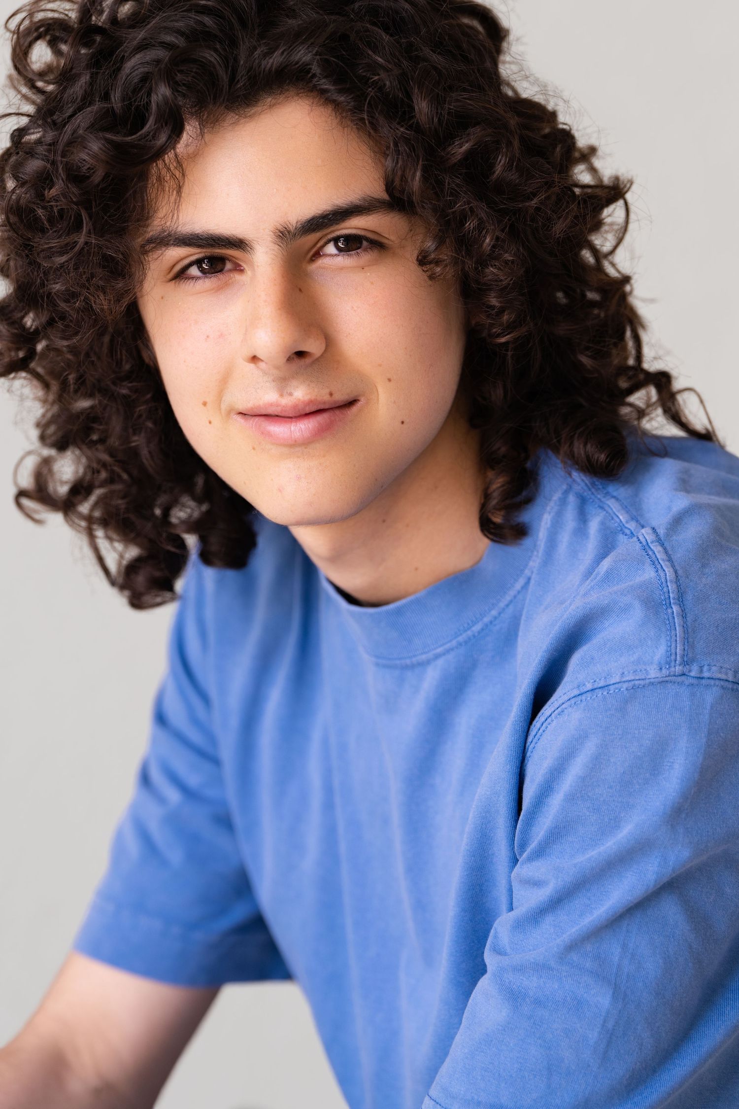 Young man with long curly dark hair wearing a blue t-shirt, smiling against a light gray background.