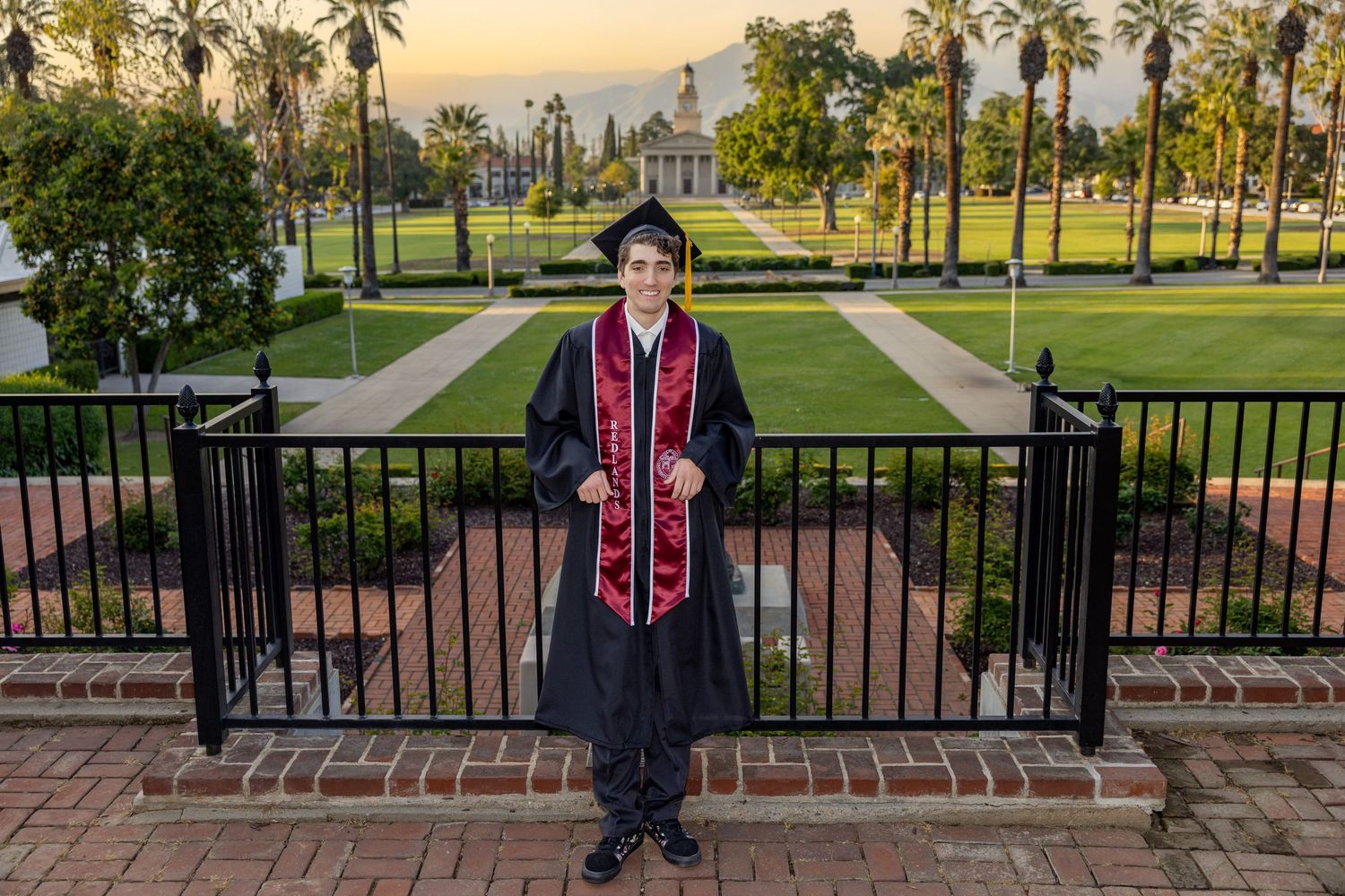 A person in graduation attire stands in front of palm trees and brick walls during golden hour at a university campus.