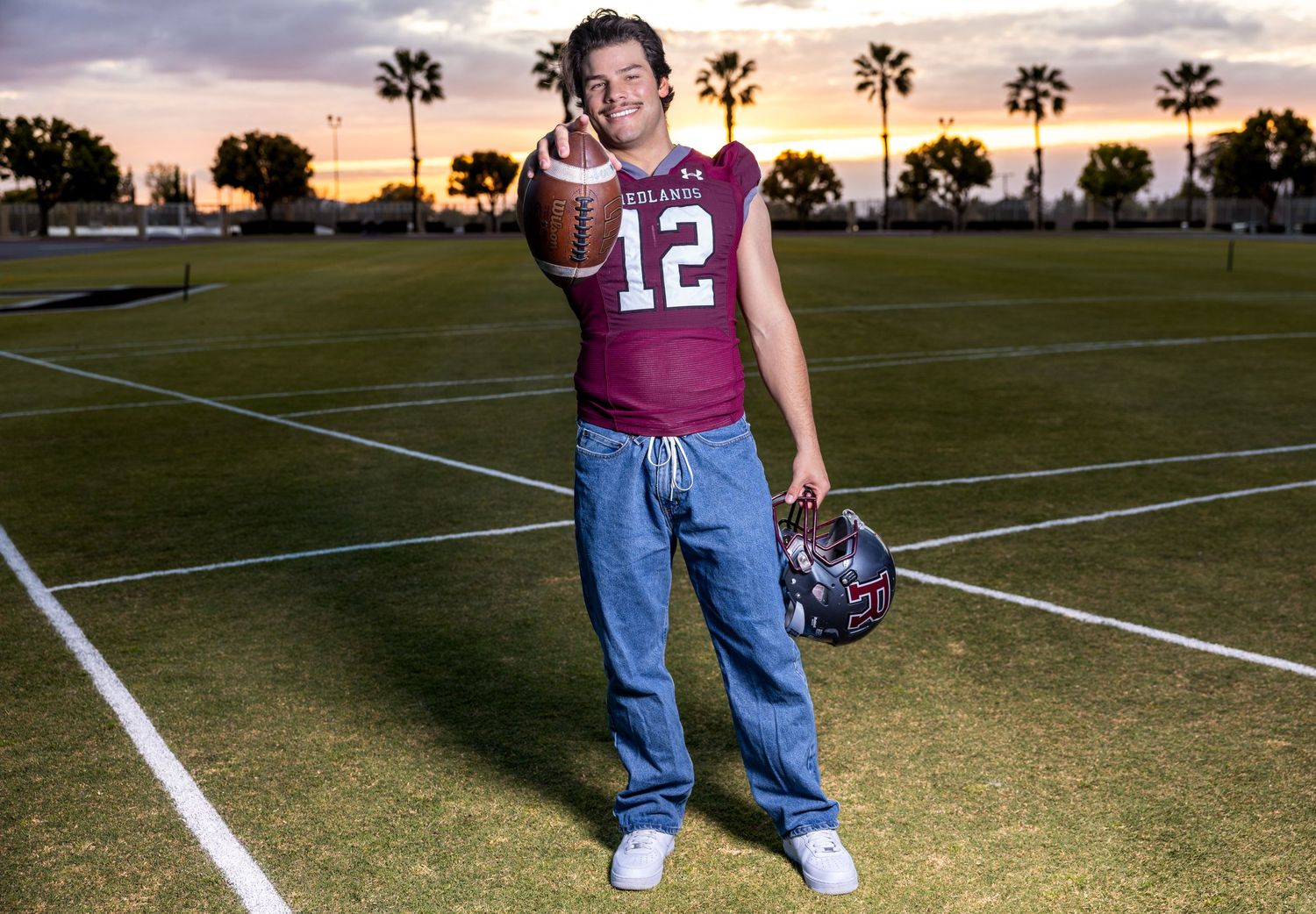 A football player stands on a field at sunset with palm trees silhouetted in the background.