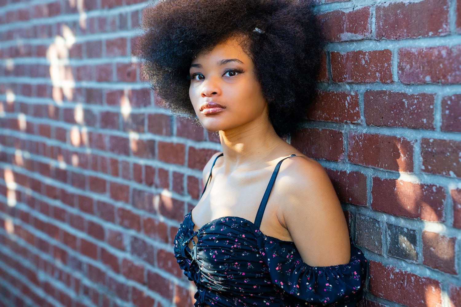 A person in a black dress poses against a brick wall with soft lighting and an urban backdrop.