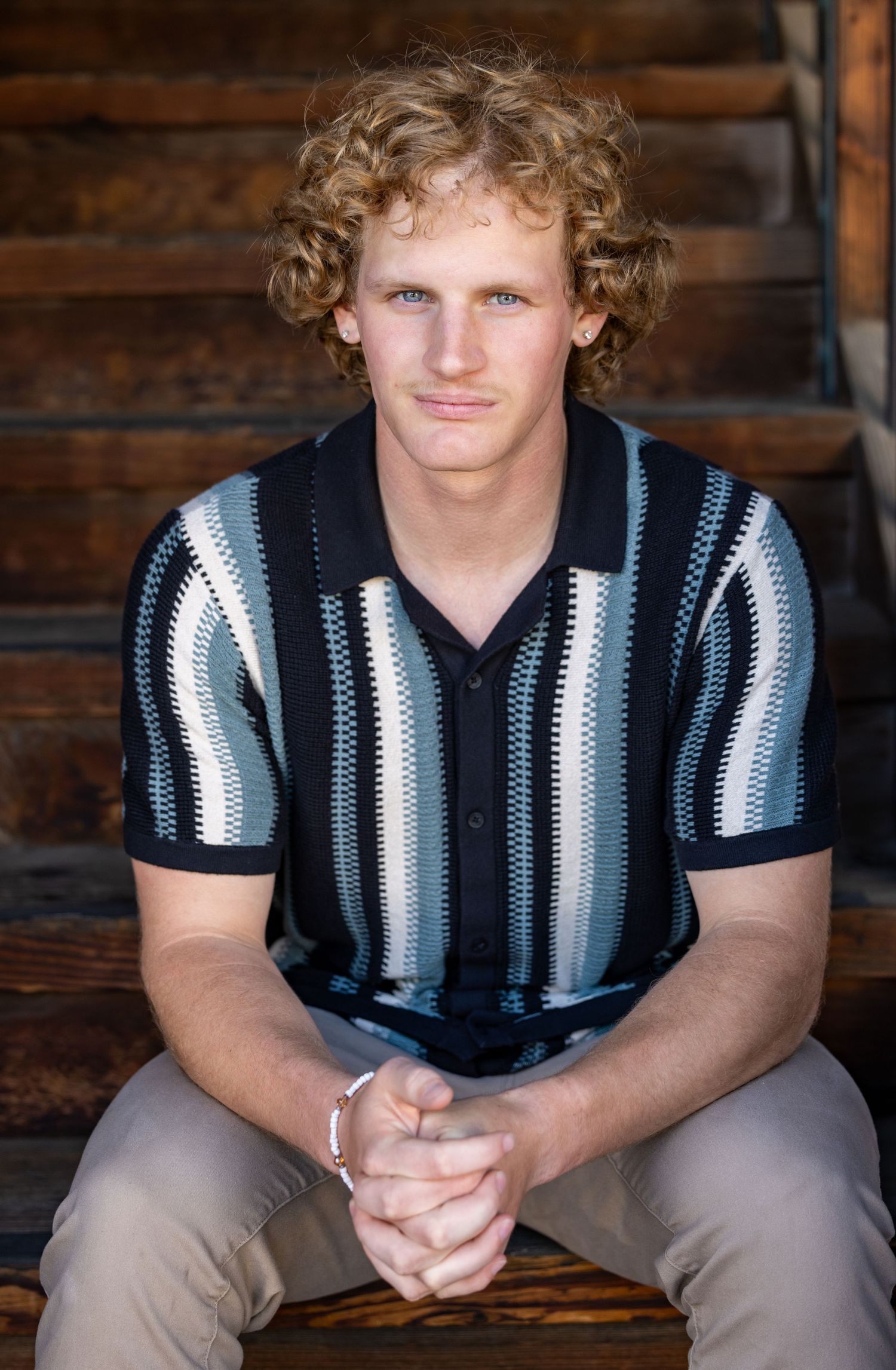 A person in a striped black and white polo shirt sits on a wooden bench against a rustic wood wall.