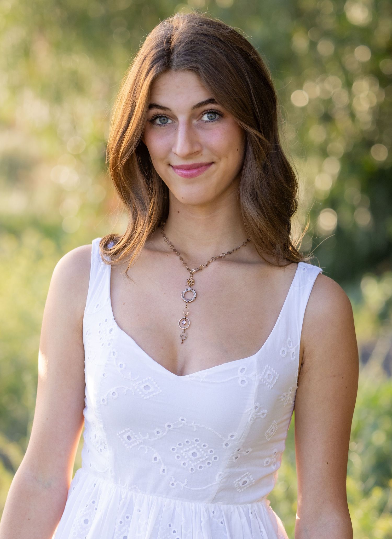 A woman in a white eyelet sundress poses in a natural outdoor setting with soft bokeh background lighting.