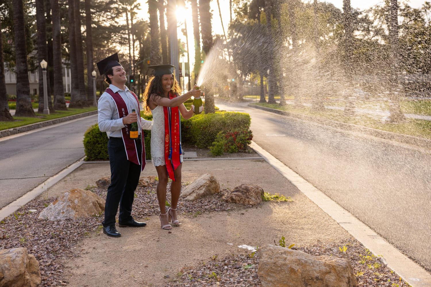 People walking through sprinklers on a sunny sidewalk while water sprays in multiple directions on a warm day.