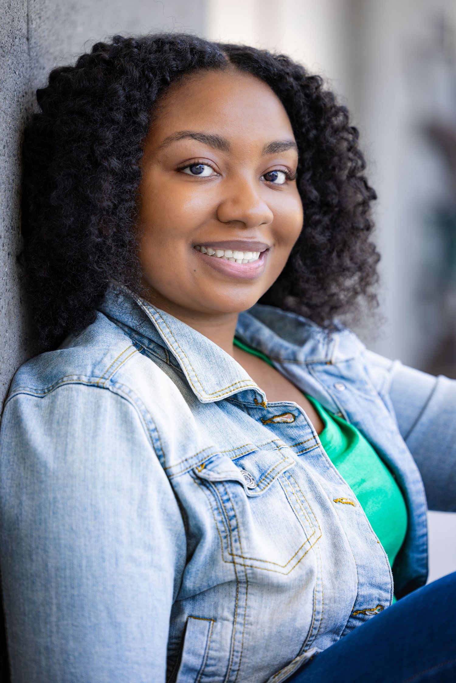 Smiling young Black woman in denim jacket and green shirt leaning against a wall outdoors.