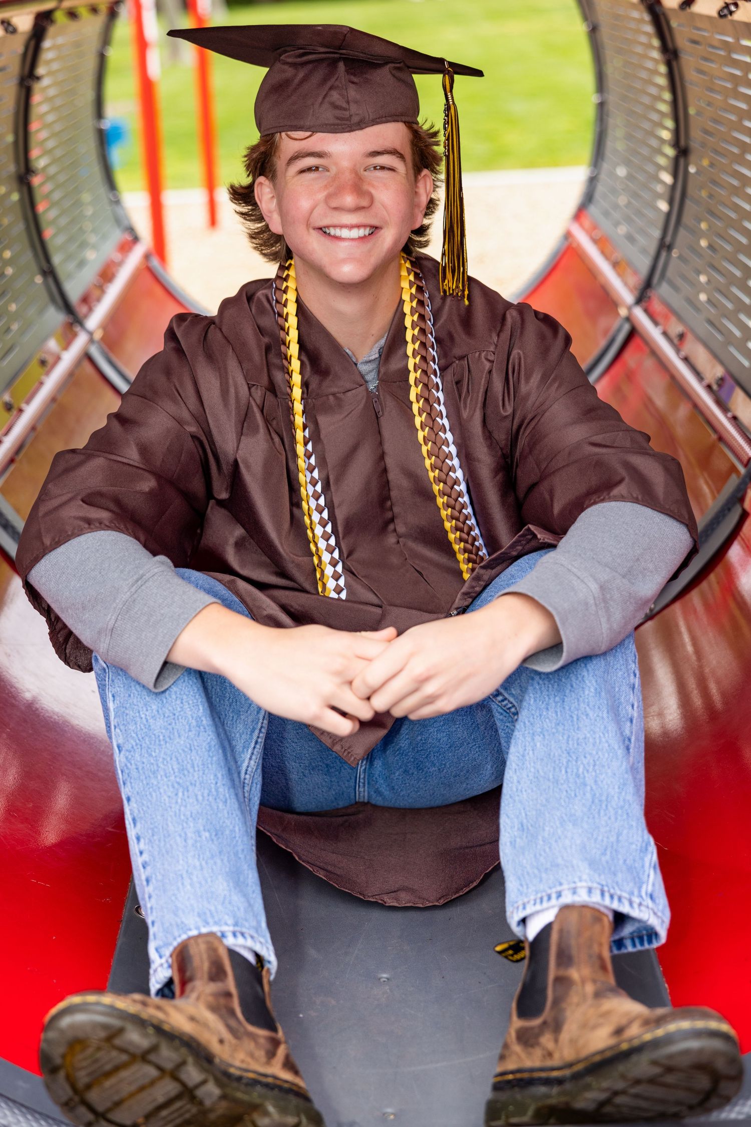 Graduate in brown cap and gown sits inside a colorful playground tunnel wearing honor cords and blue jeans.