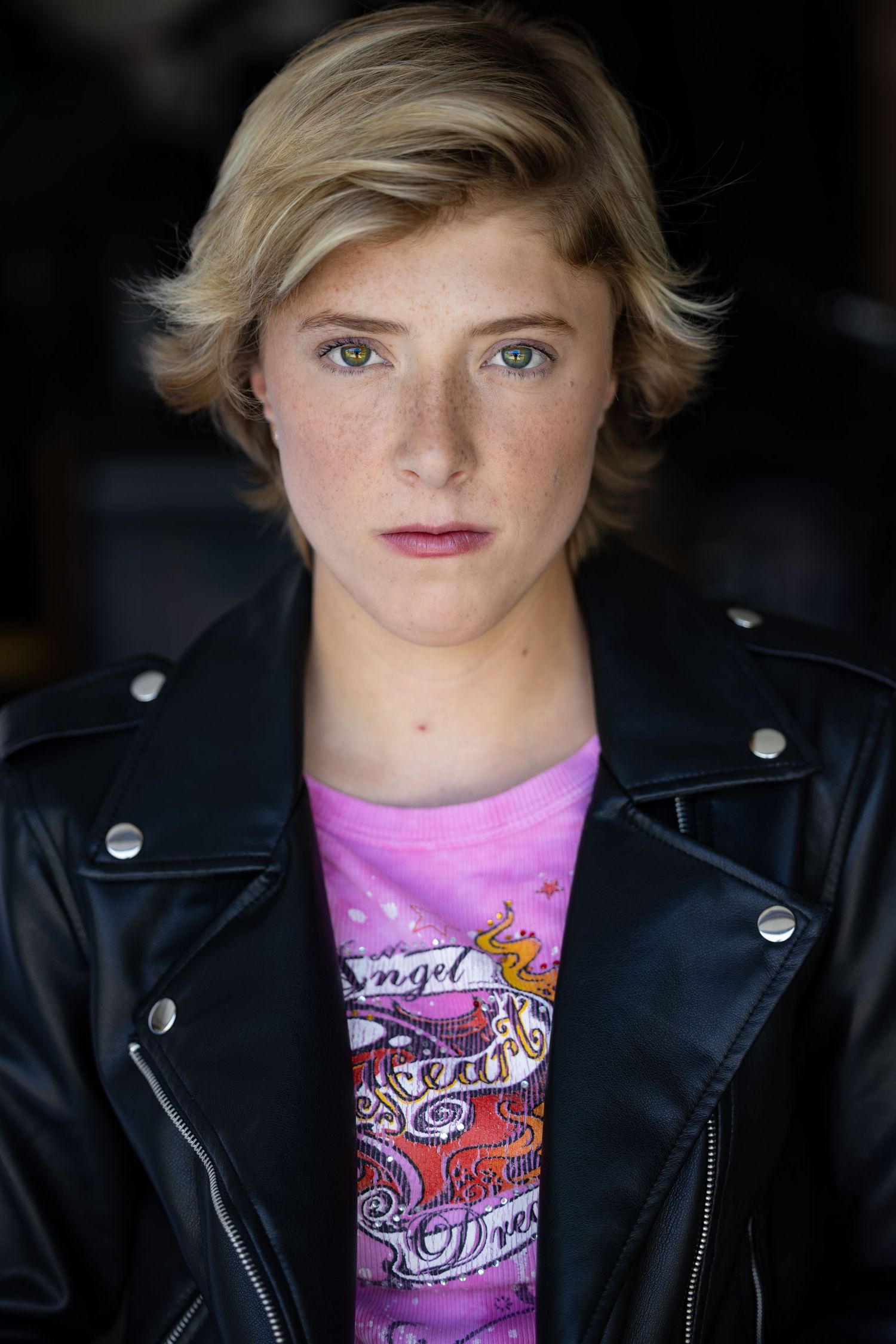 theatrical headshot. punk girl. Portrait against dark background wearing black leather jacket and pink graphic t-shirt.