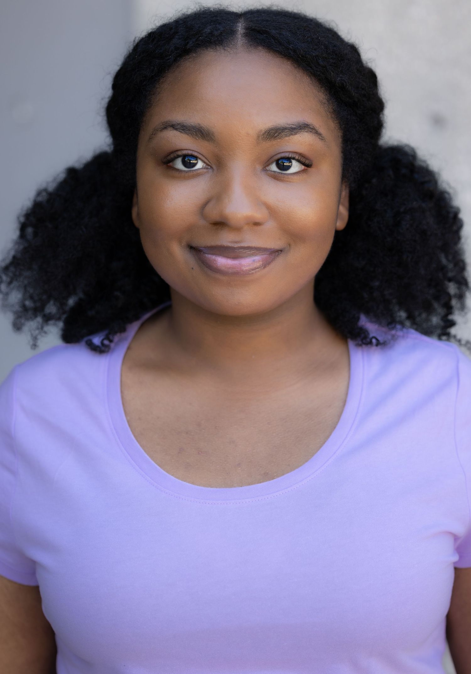 Portrait series in lavender top showing warm smile and natural curly hair against light background.