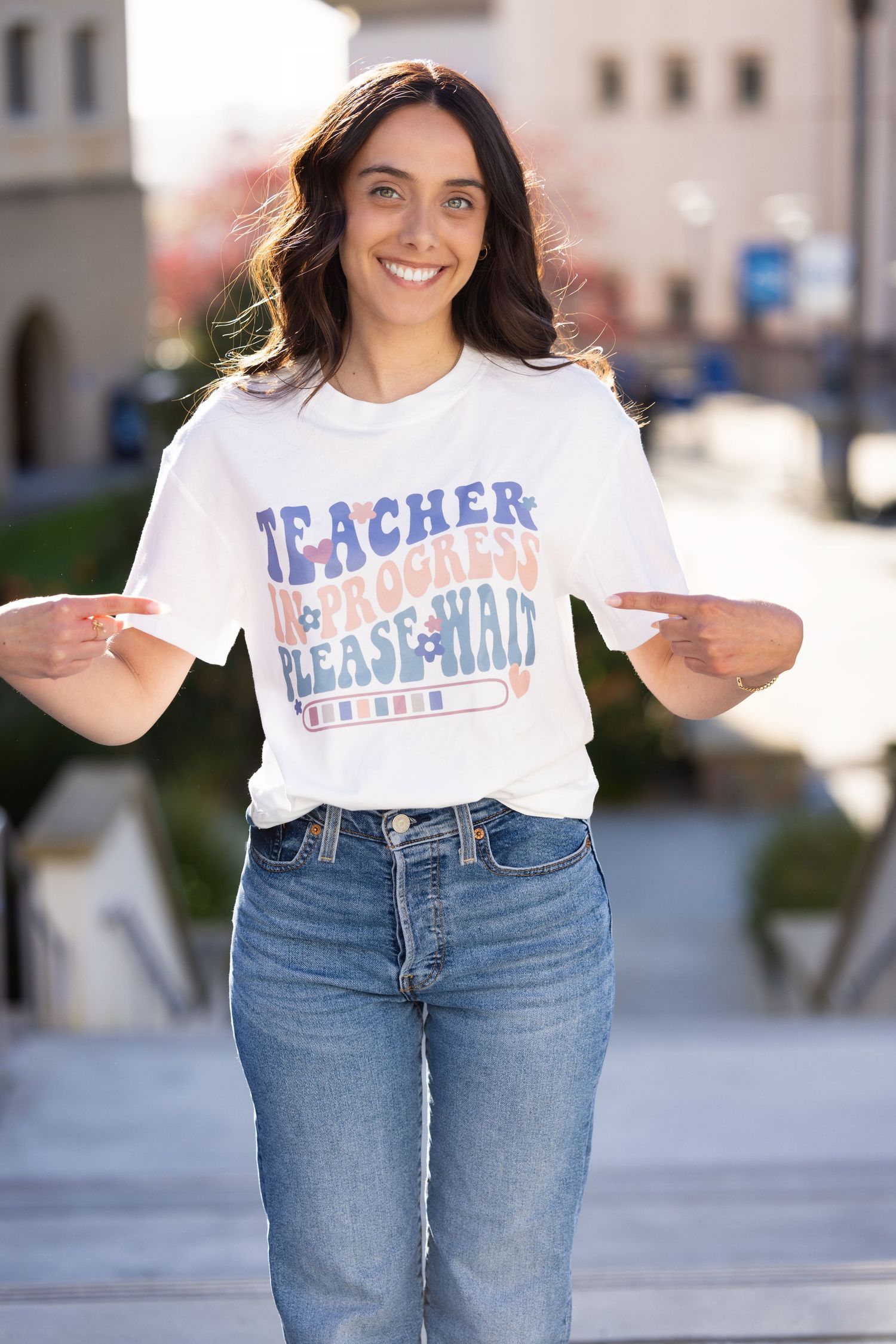Person wearing a white t-shirt with 'Teacher Baseball' text and blue jeans poses outdoors with a bright smile.