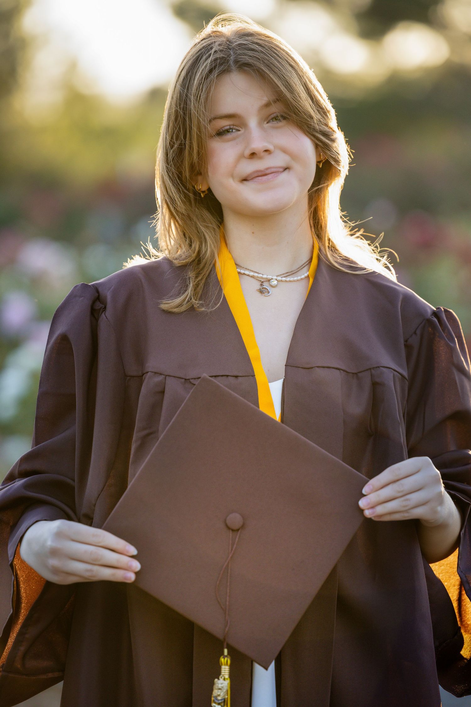 Graduate in brown academic regalia and yellow stole smiles while adjusting their gown during commencement ceremony.