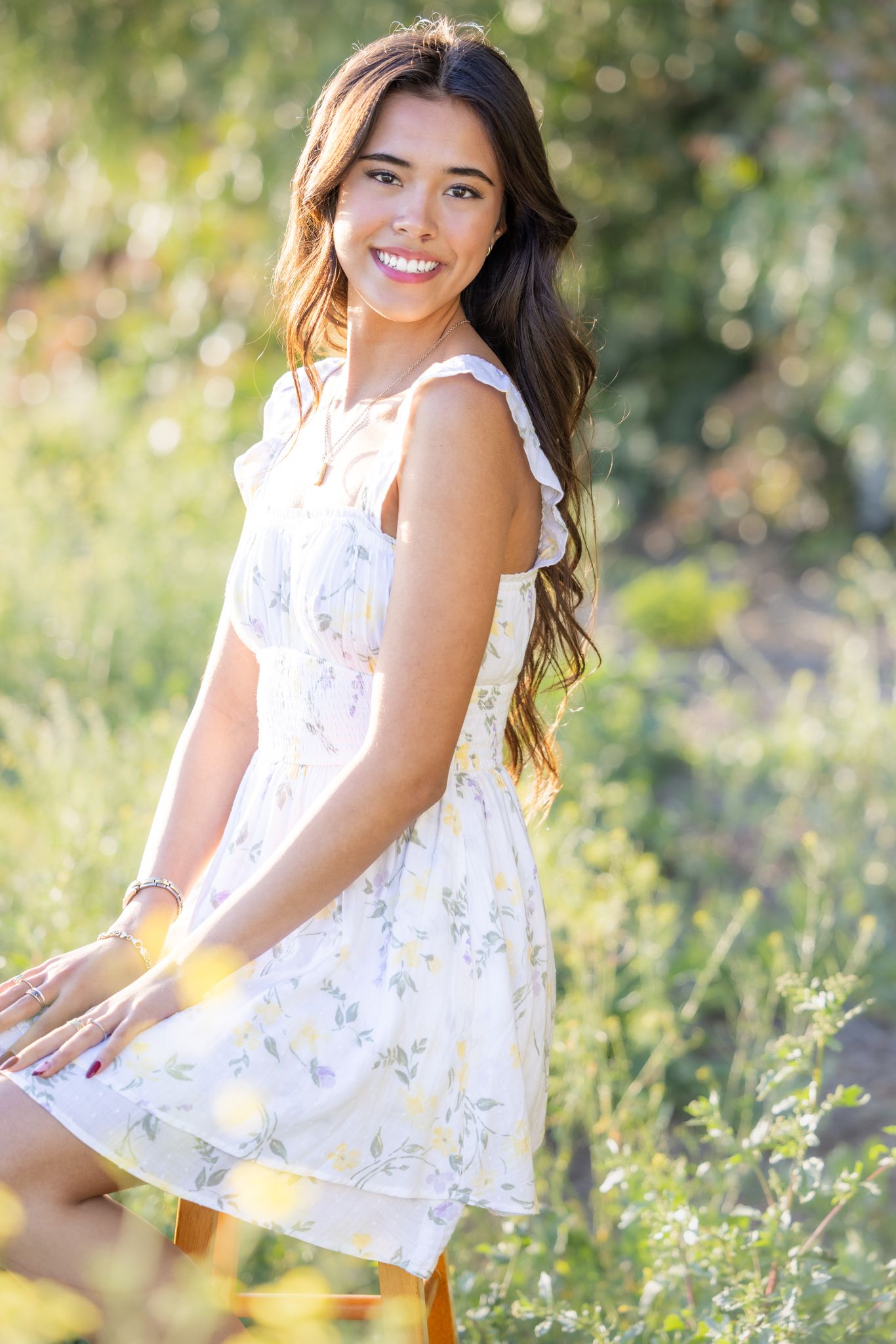 A person in a white floral dress sits in a sunny meadow with soft natural lighting and green foliage in the background.
