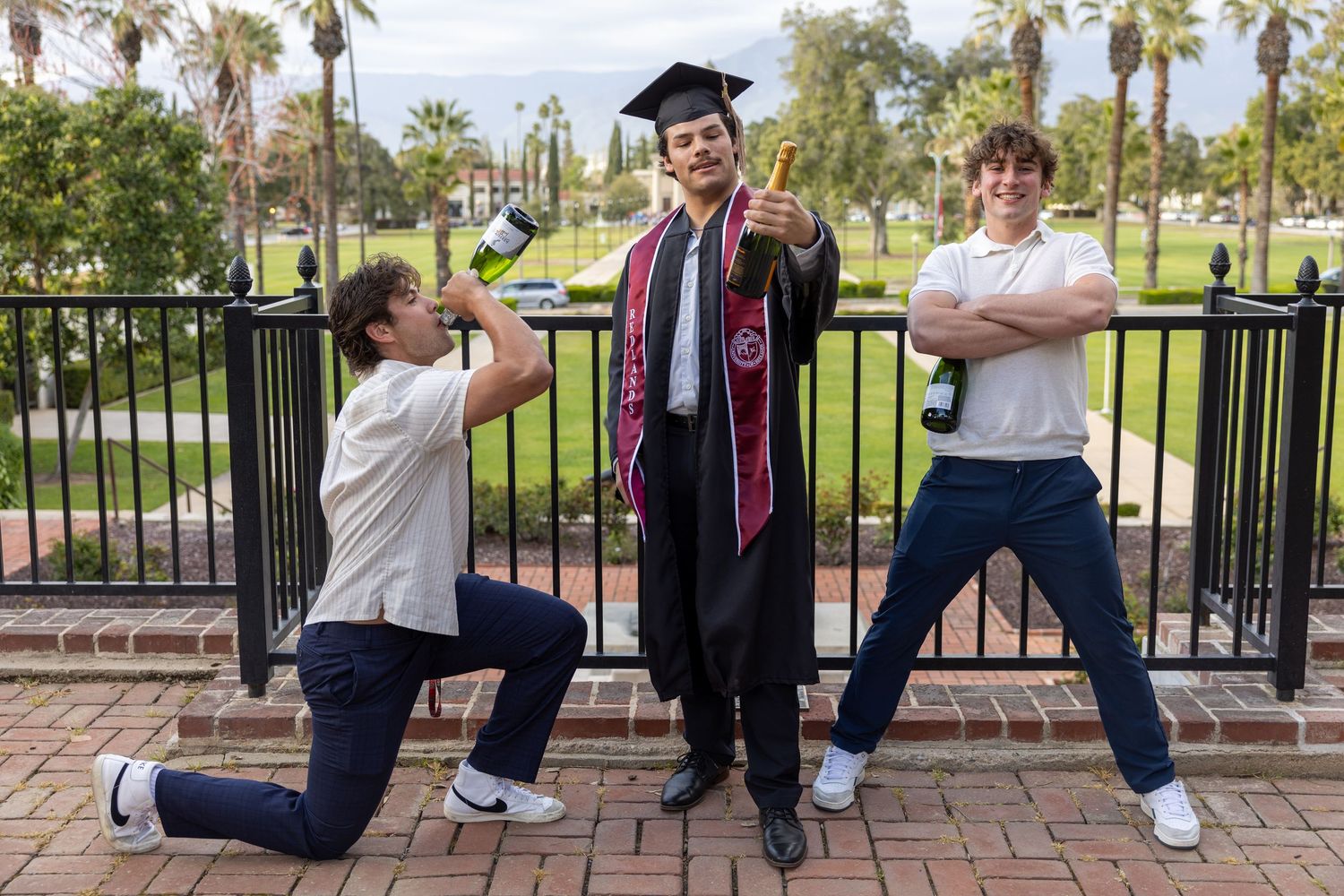 Three people pose playfully on a brick path with palm trees, one wearing graduation regalia with cap and gown.