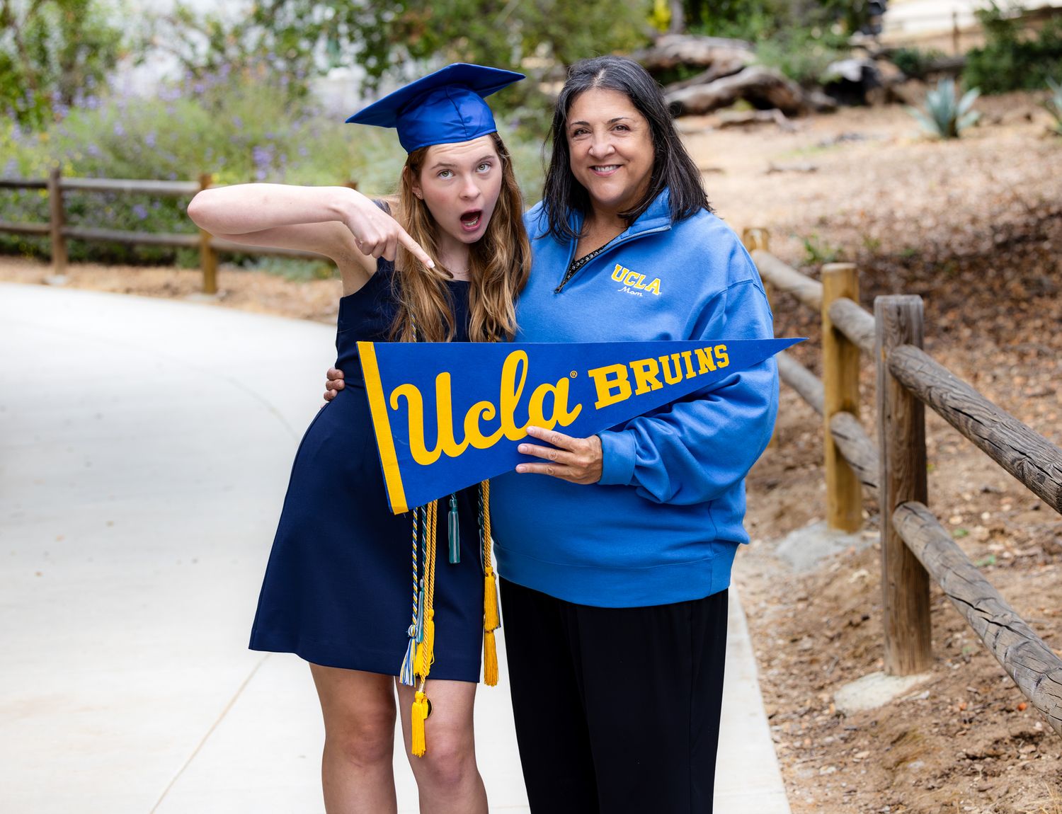 Two people in UCLA graduation attire pose together on a wooden path during a sunny day.
