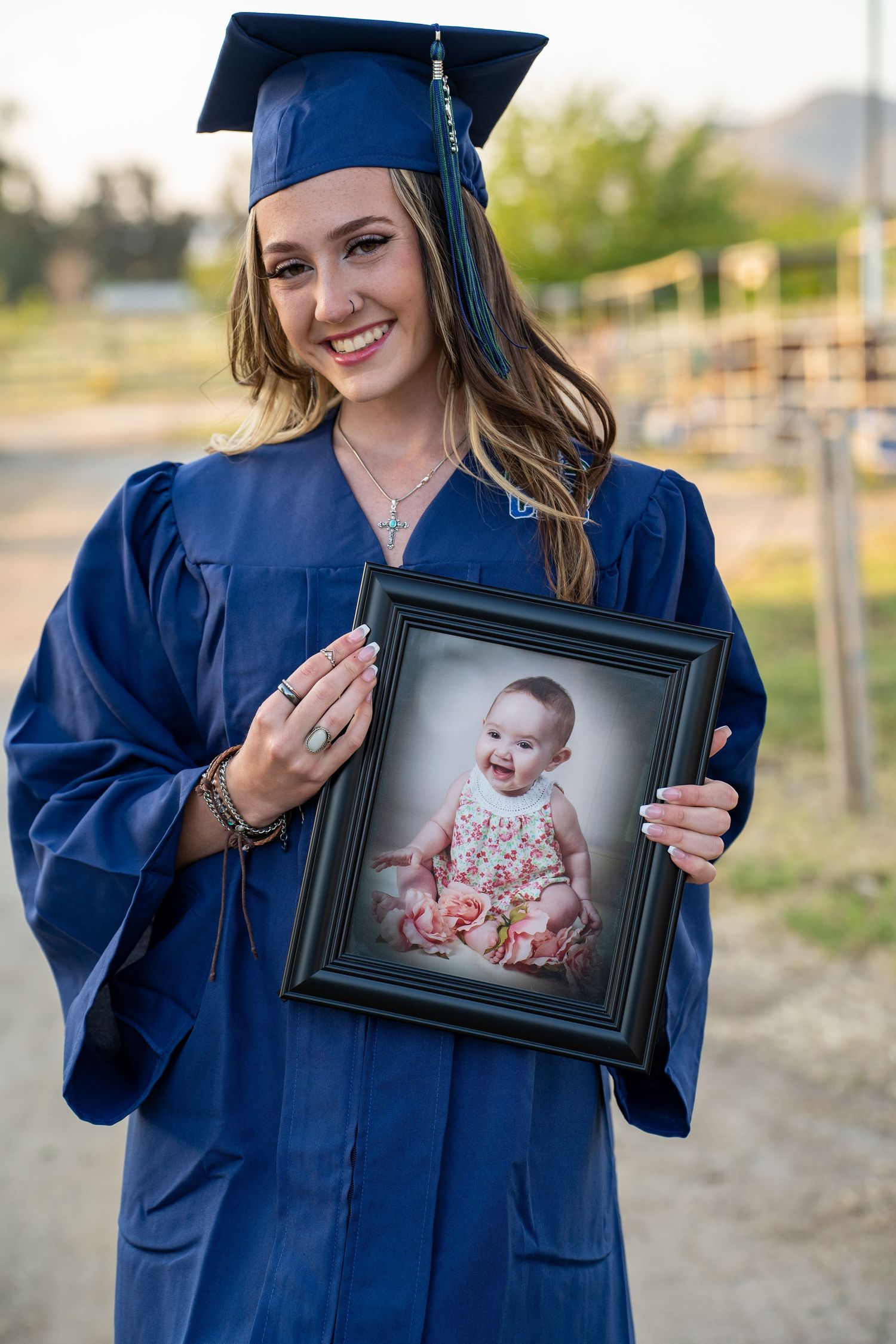 Graduate in navy blue cap and gown holds framed baby photo during outdoor photoshoot.