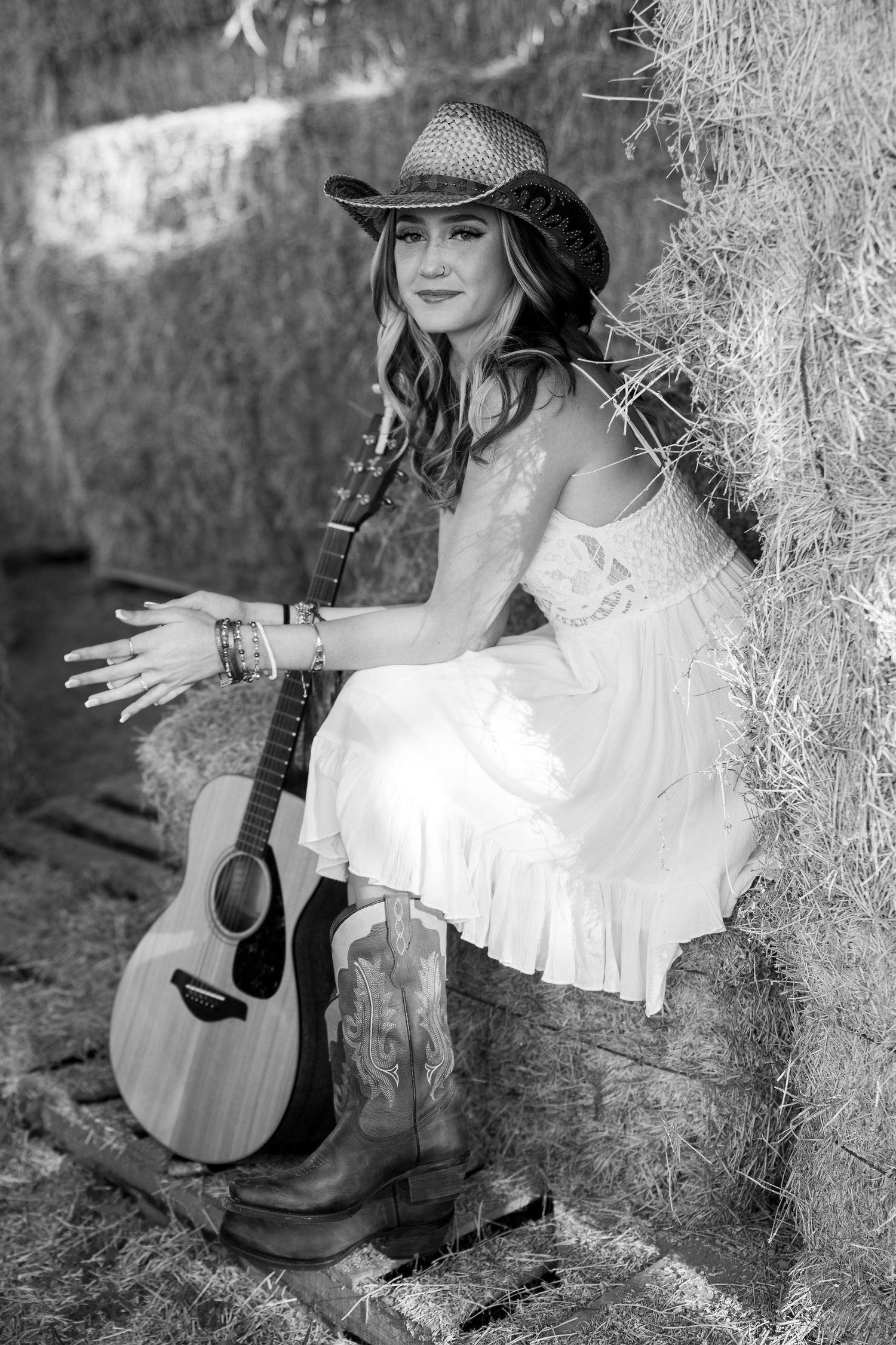 Black and white country western portrait with acoustic guitar and cowboy boots against rustic hay bale backdrop.