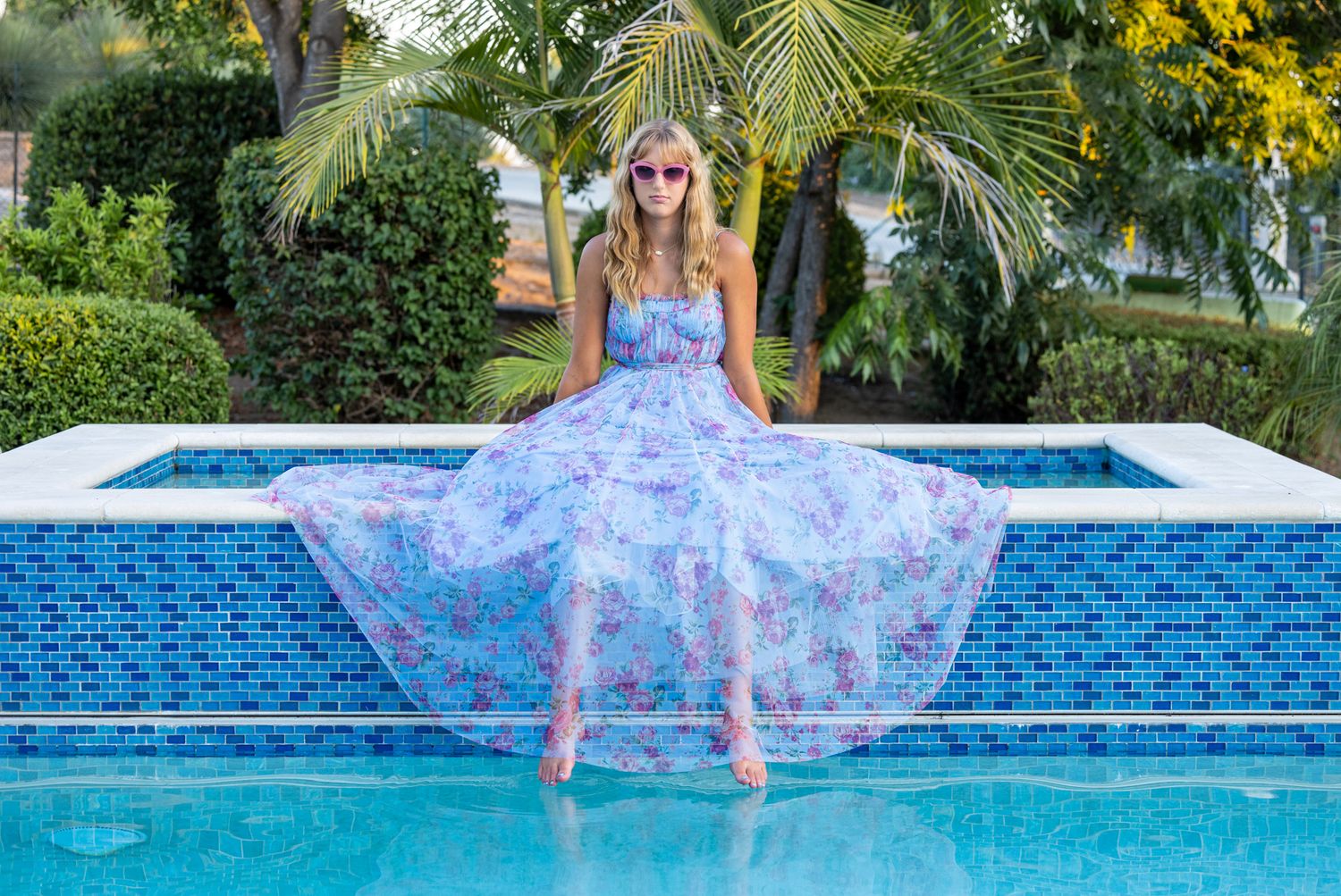 A flowing floral dress billows gracefully over a bright blue swimming pool on a sunny tropical day.