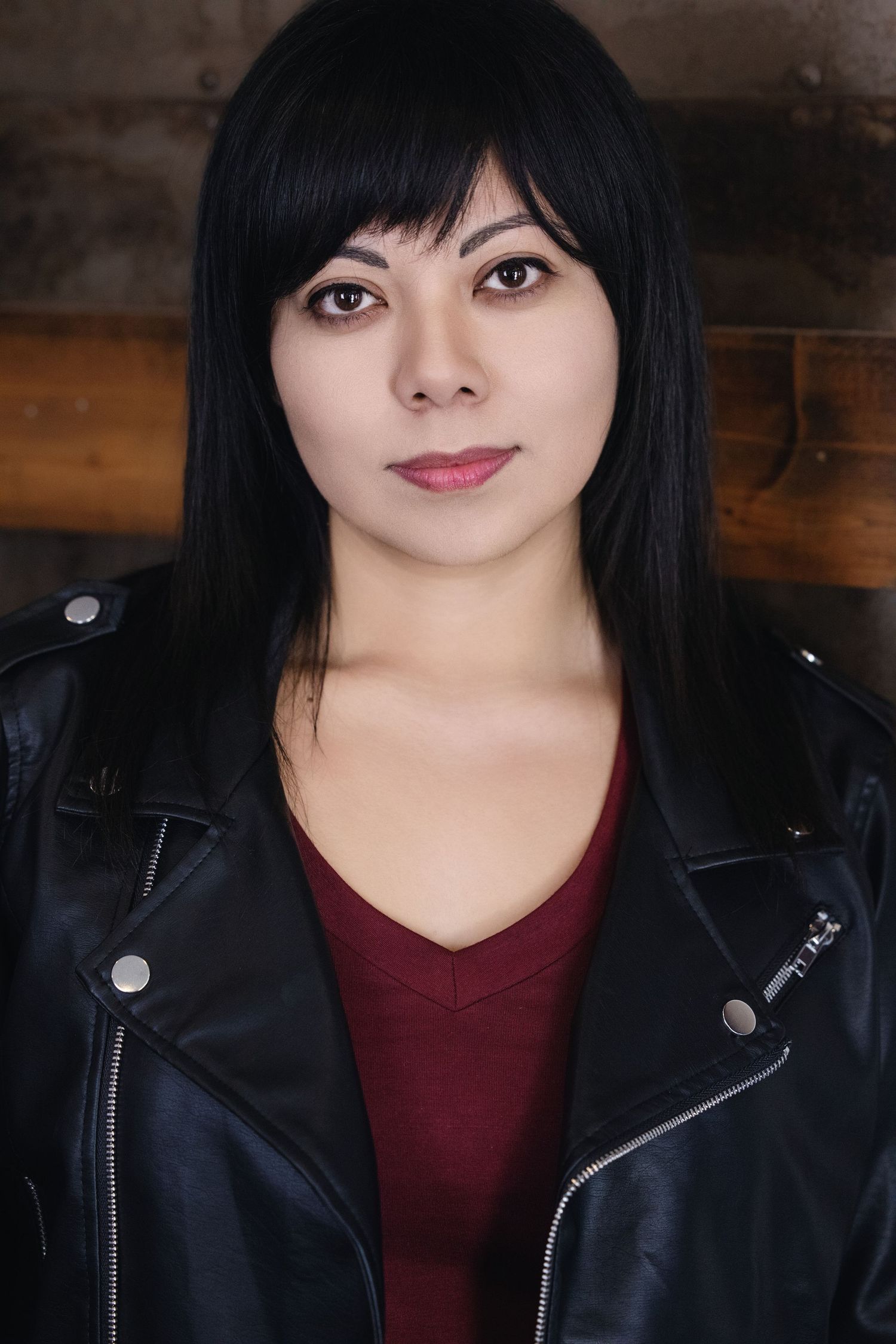Close up fashion headshot of someone wearing a black leather jacket and burgundy top with dark bangs against wooden backdrop. detective girl