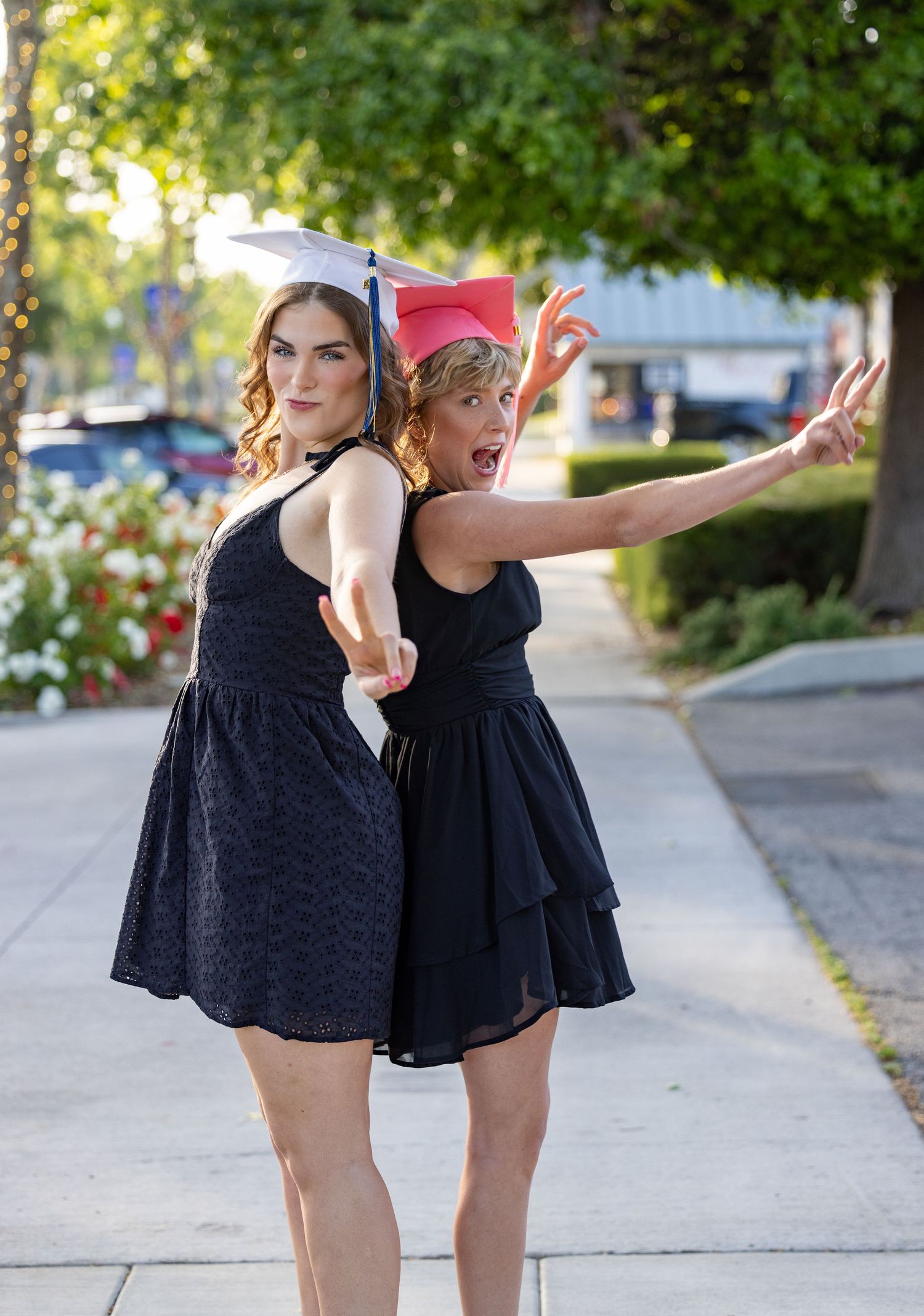 Two women in black dresses strike playful poses together on a sunny sidewalk with trees in the background.