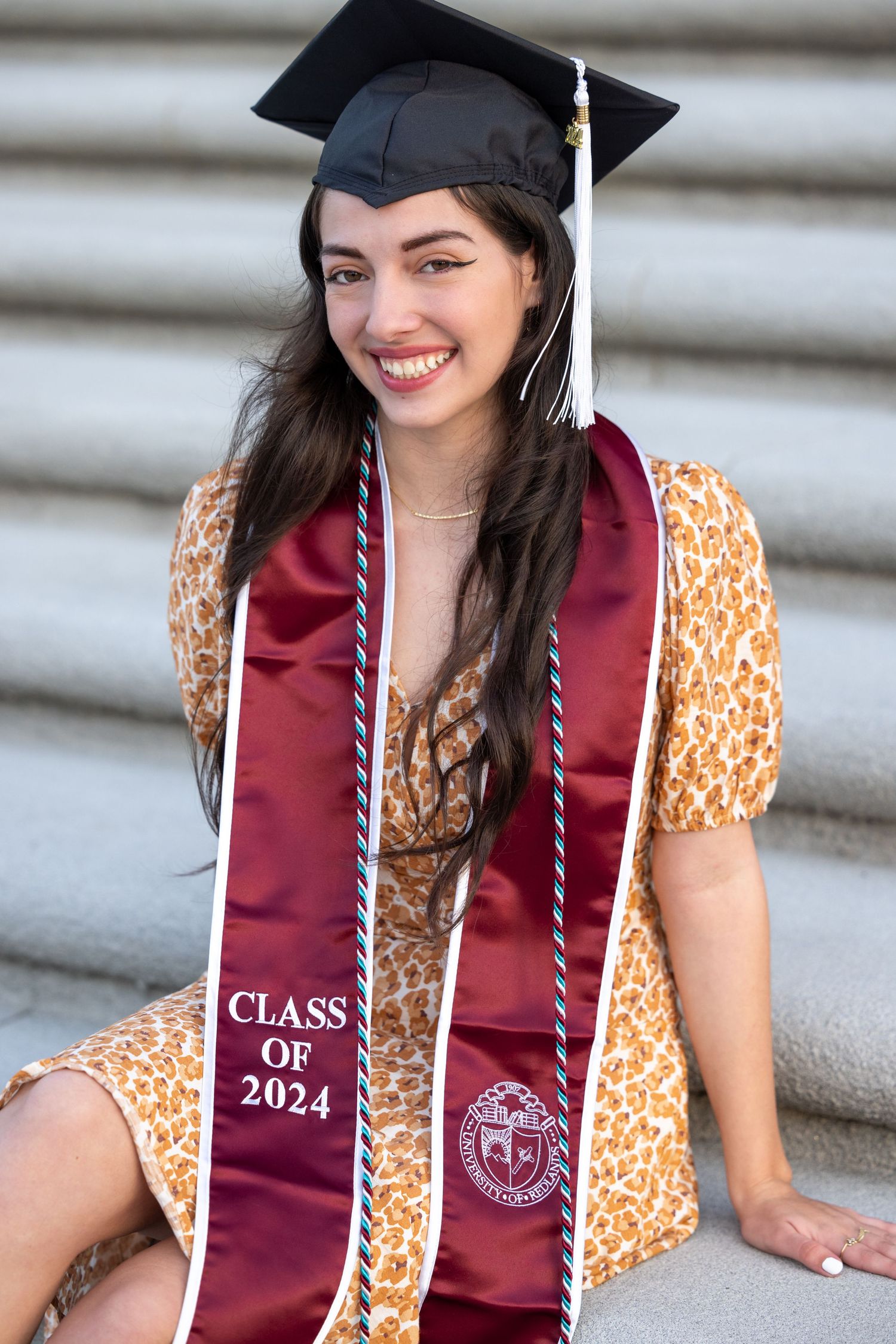 A graduate in a burgundy stole and orange floral dress poses for graduation photos on stone steps.