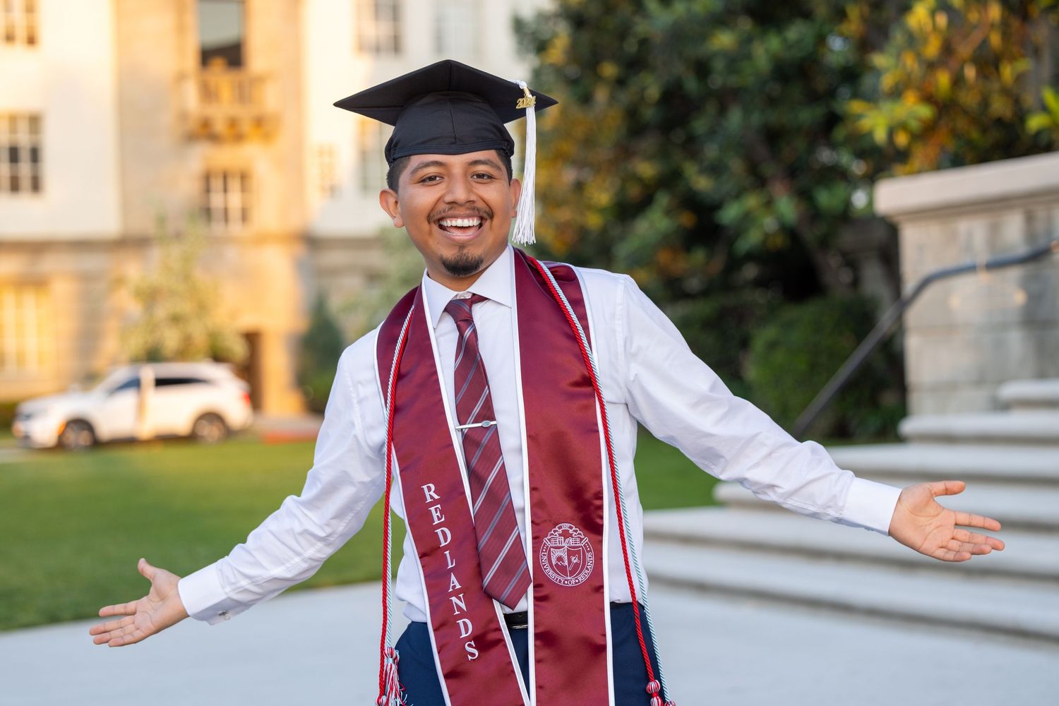 Graduate in burgundy vest and graduation cap poses on outdoor campus steps during golden hour.