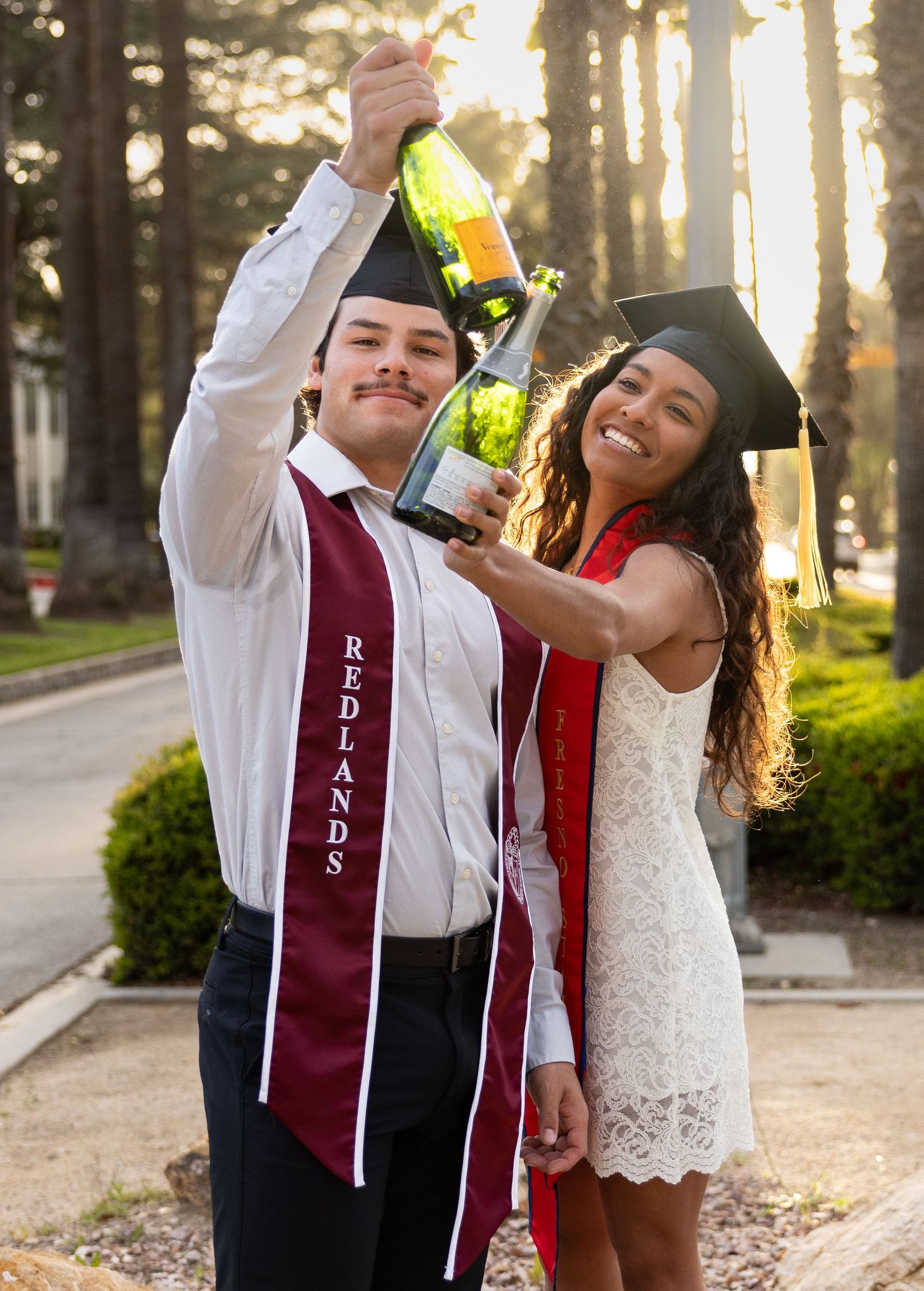 A couple in graduation attire sharing a celebratory drink at sunset in a wooded outdoor setting.