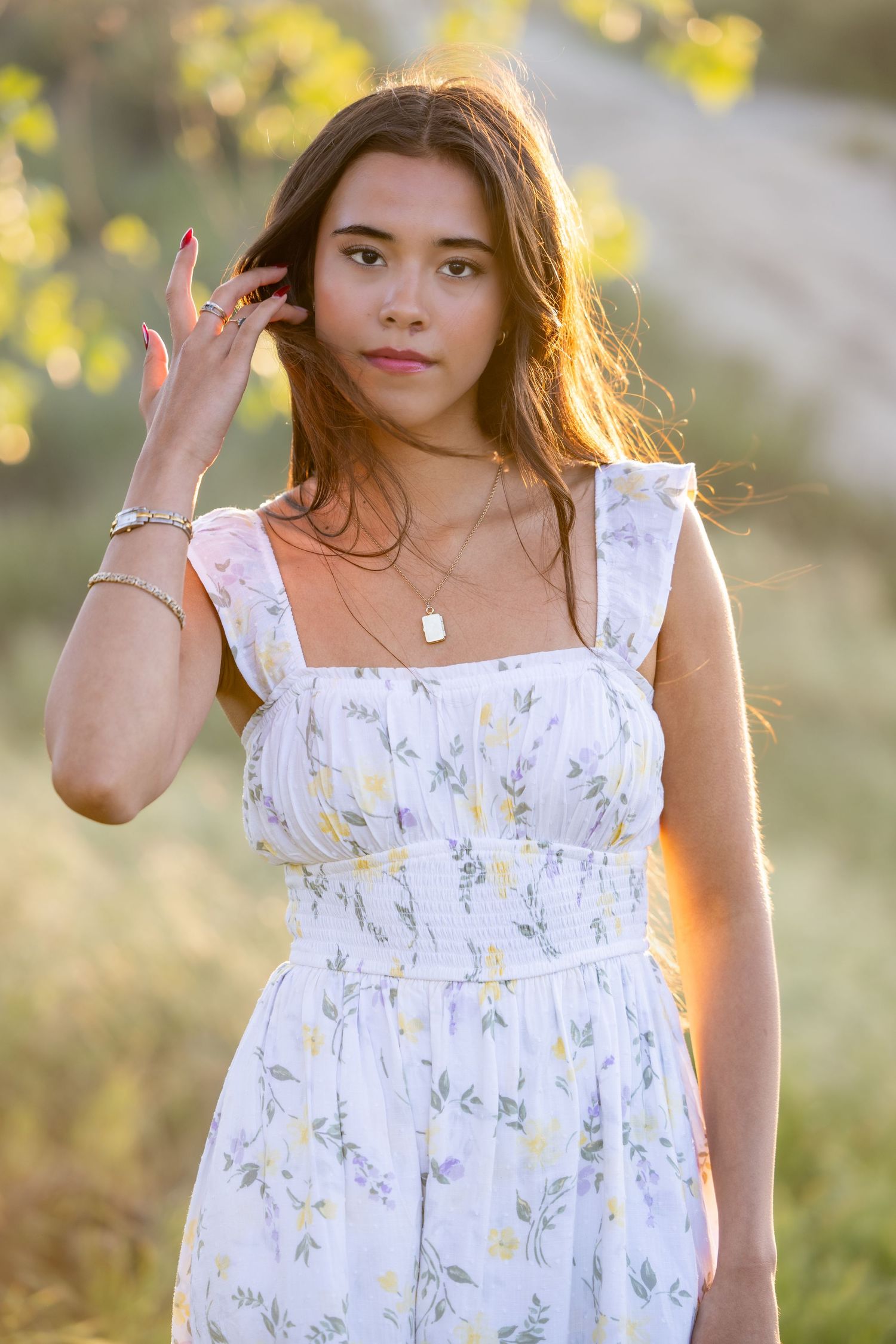 A person in a white floral sundress poses outdoors during golden hour with sunlit foliage in the background.