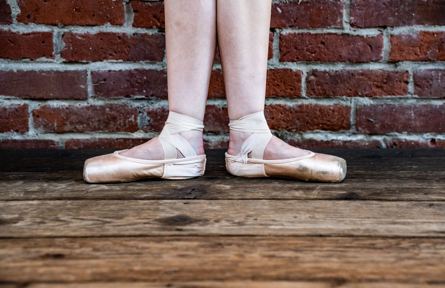 Two pairs of feet in ballet shoes stand against a rustic brick wall background.