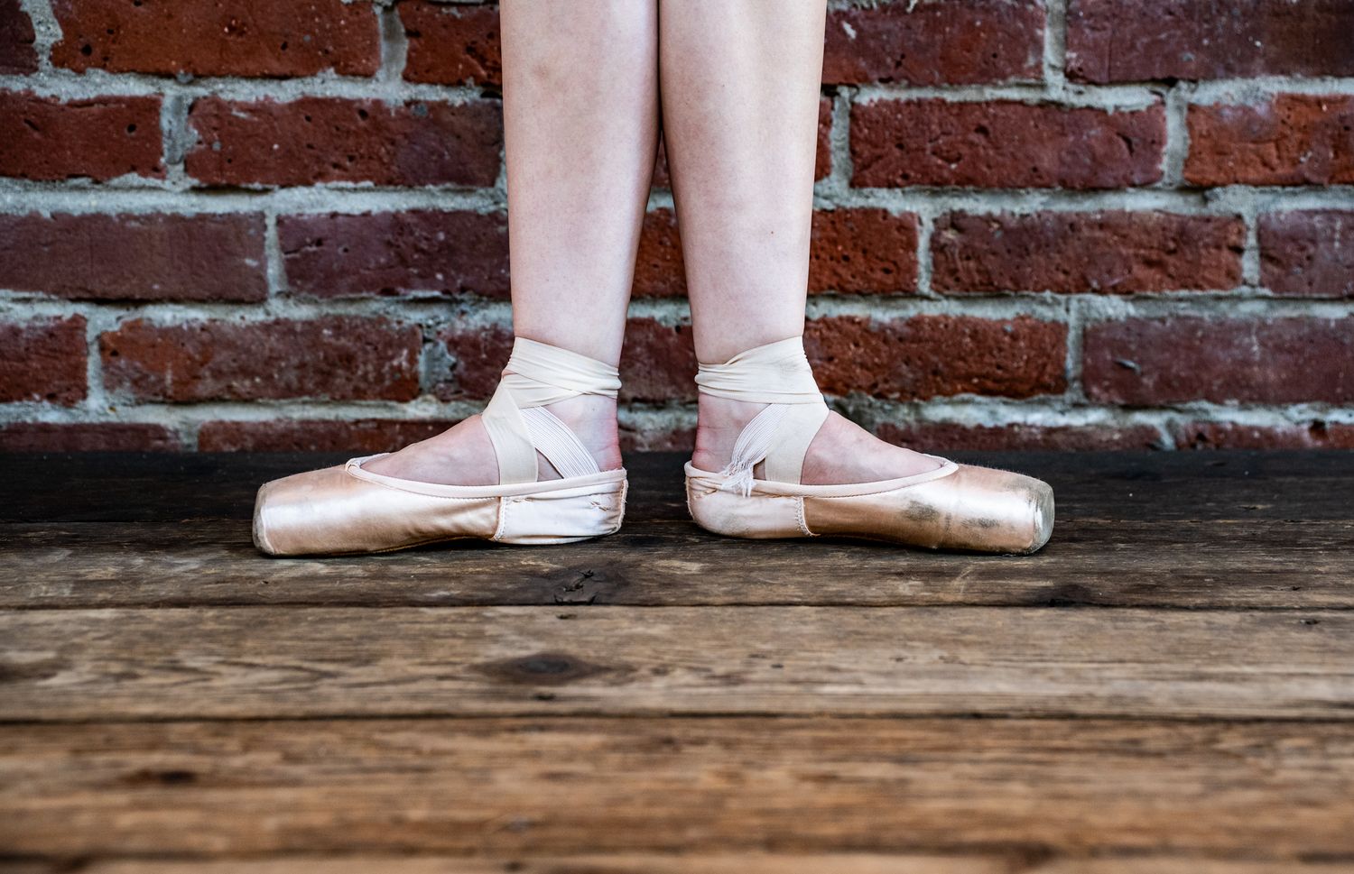 Ballet pointe shoes and feet photographed against a rustic brick wall and wooden floor.