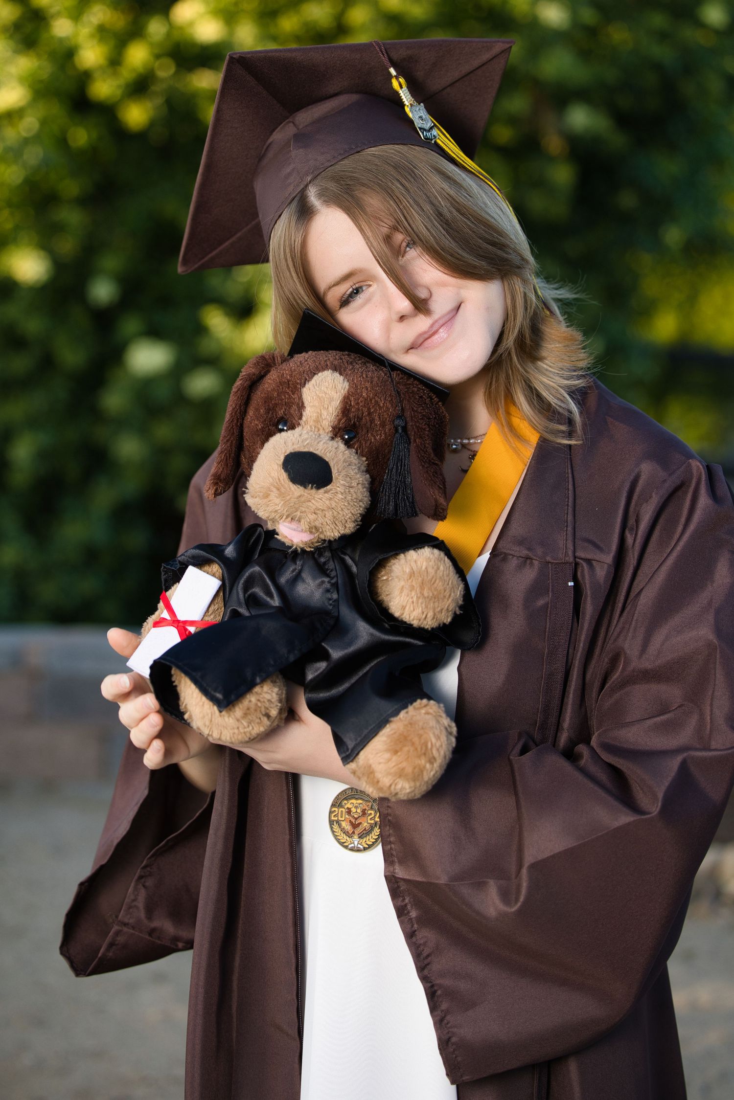 Graduate in brown cap and gown holds stuffed puppy dog wearing matching graduation attire during commencement ceremony.