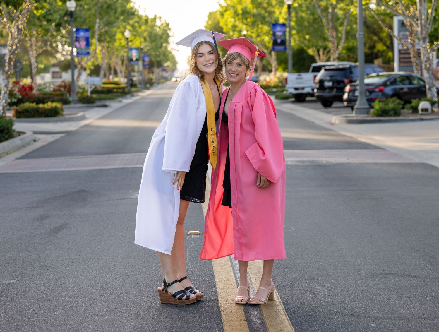 Two people in graduation attire walk down a suburban street on a sunny autumn day.