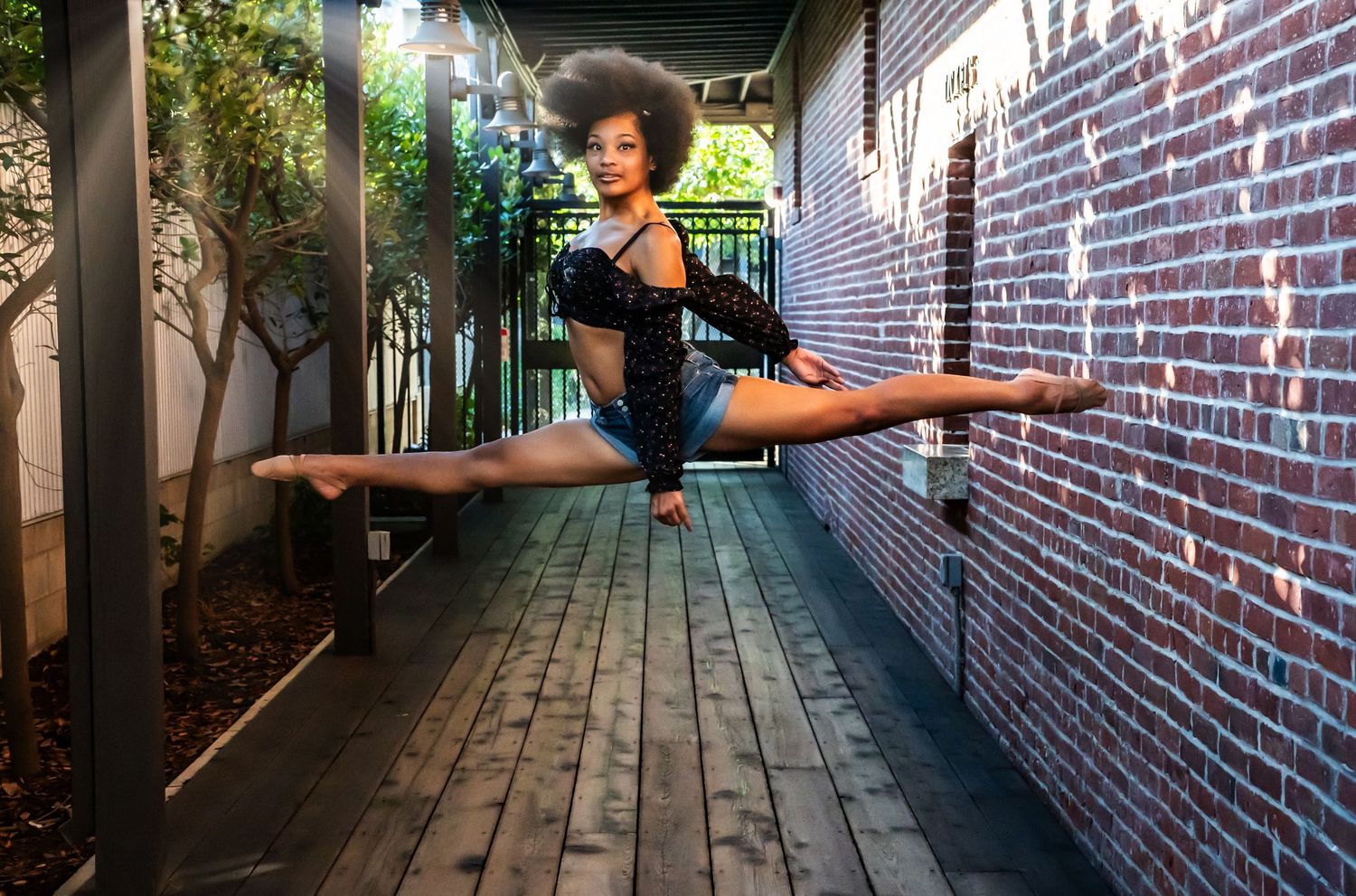 A dancer practices ballet poses in a sunlit brick corridor with dramatic shadows.
