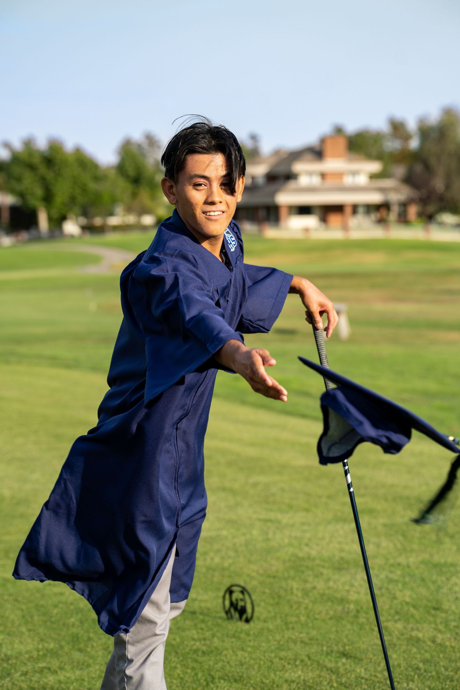 A child in a navy blue jacket plays outdoors on a grassy field with a golf club on a sunny day.