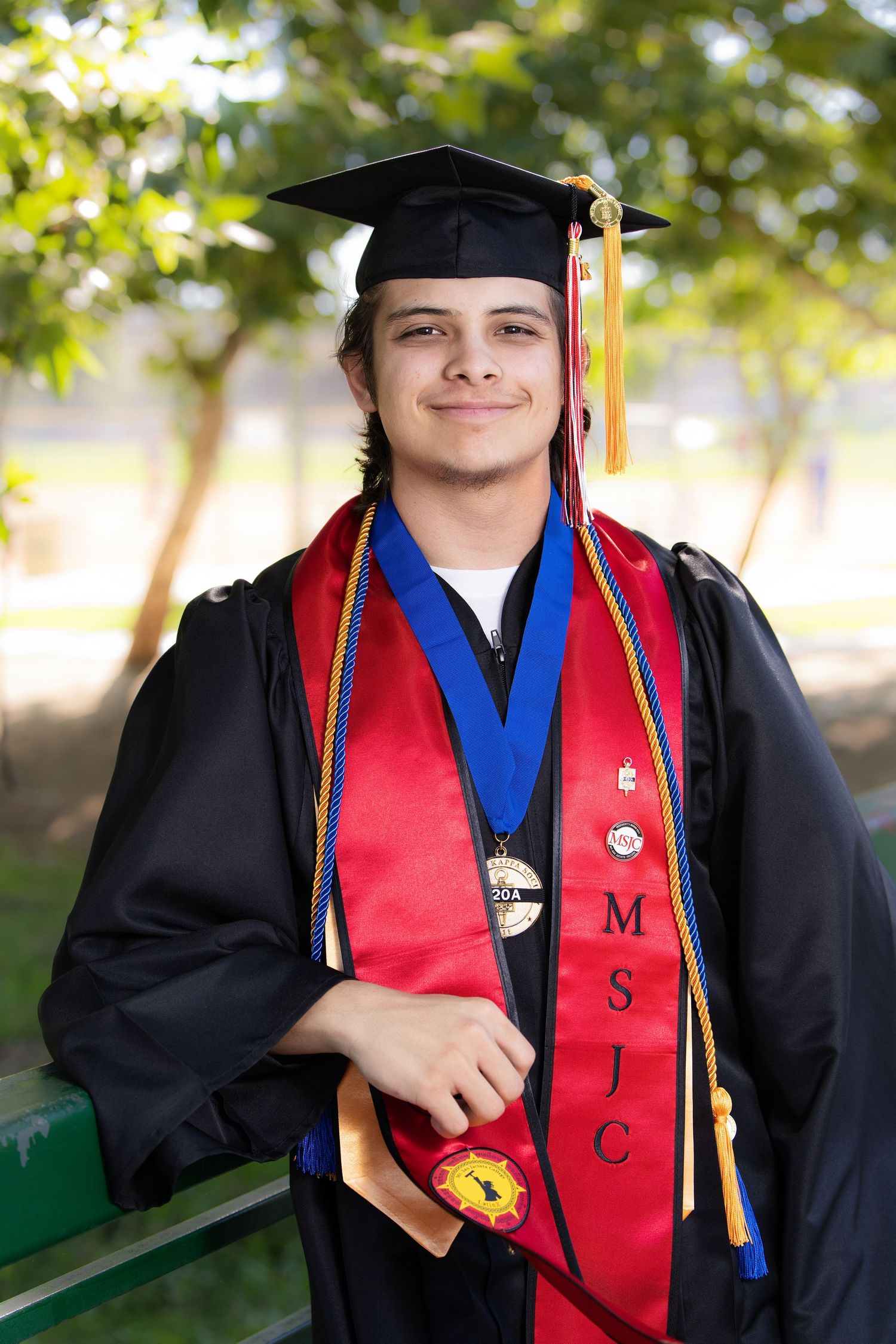 Young male graduate smiling in black cap and gown with red MSJC stole, blue cord, and medal outdoors.