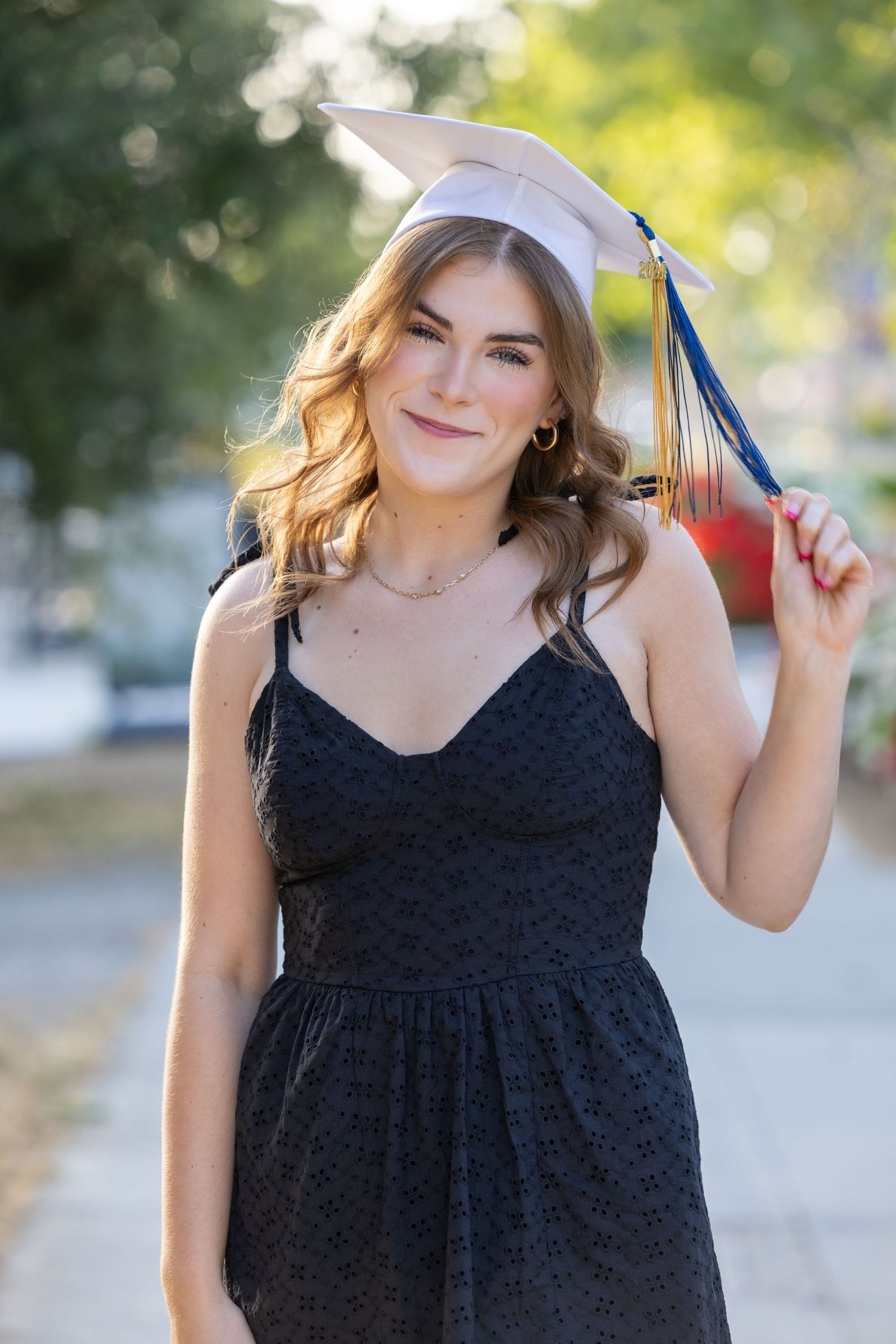 A graduate in a black dress poses outdoors with their graduation cap against a soft bokeh background.